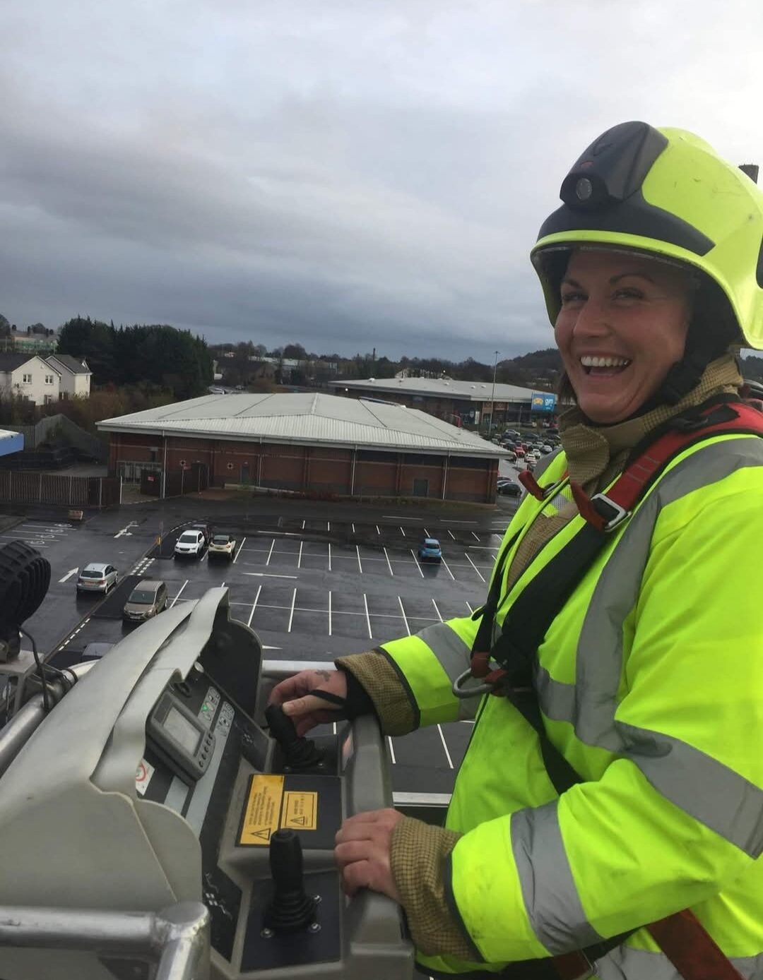 A smiling firefighter in a yellow helmet and reflective jacket stands on an elevated platform, operating controls. A parking lot and buildings are visible in the background under a cloudy sky.