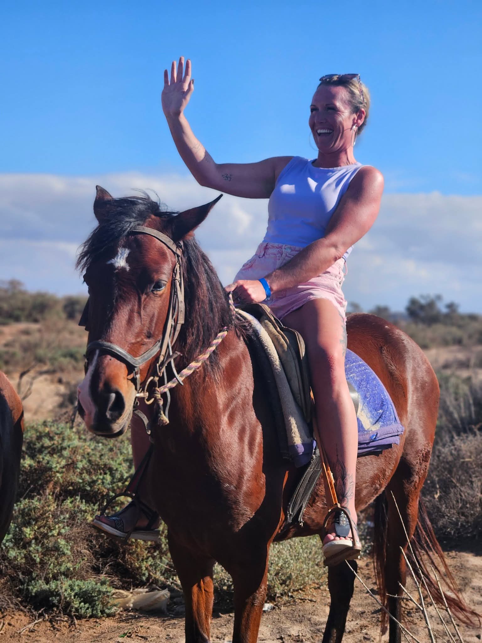 A woman wearing a light blue tank top and pink shorts is smiling and waving while sitting on a brown horse outdoors under a clear blue sky. Shrubs and dry grass are visible in the background.