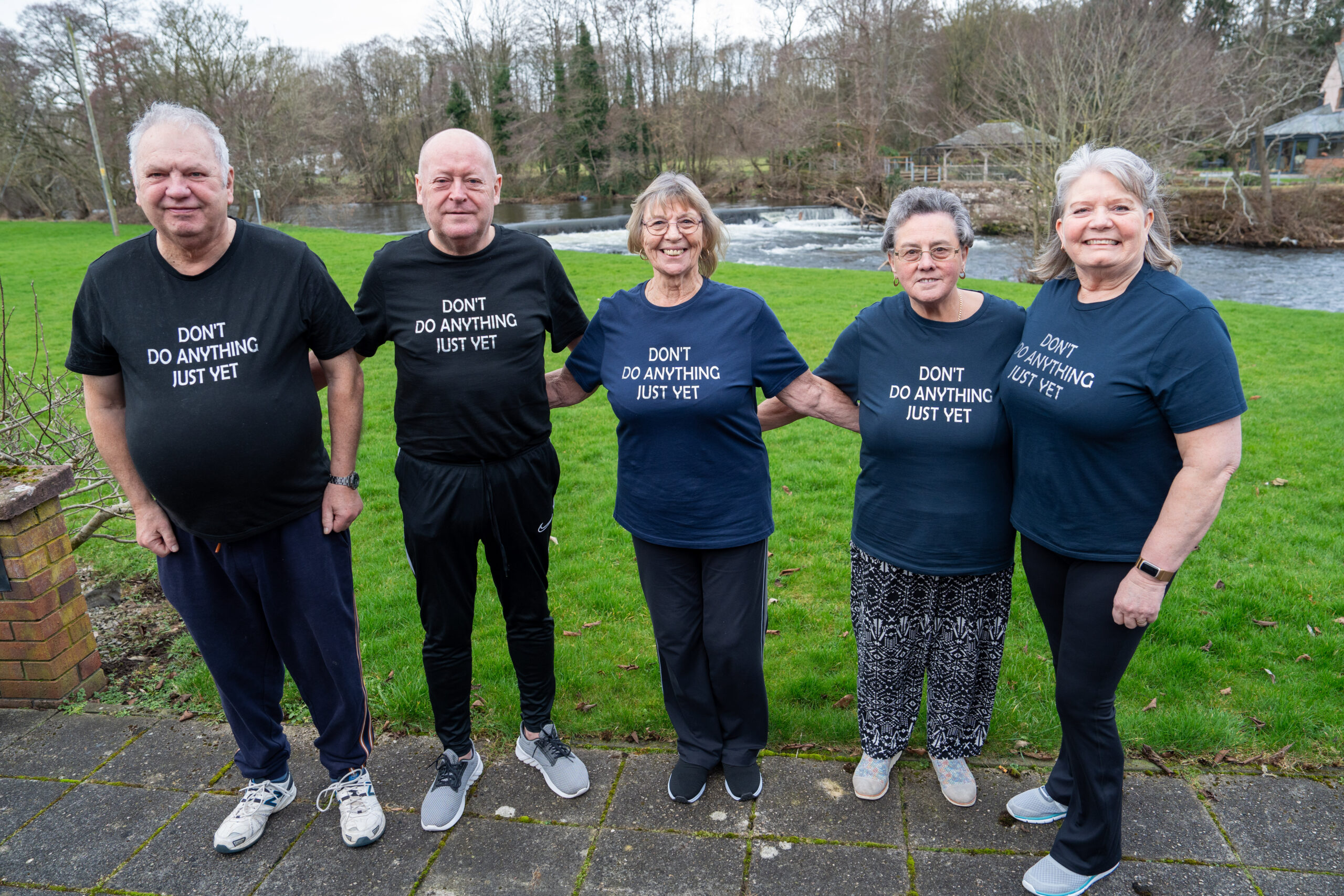 Five older adults stand outside in front of a river, smiling and wearing matching navy T-shirts that say “DON’T DO ANYTHING JUST YET.” They are standing close together on a paved path with green grass behind them.