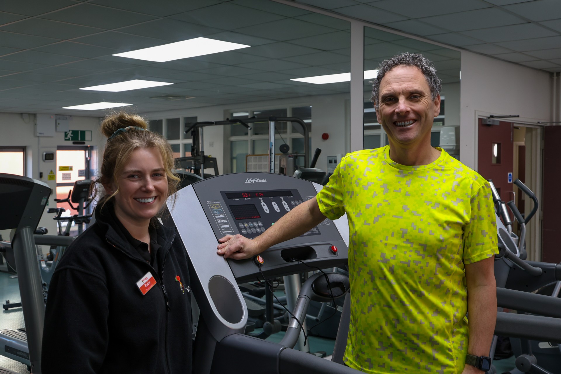 A smiling man in a bright yellow shirt stands next to a treadmill with a woman in a black jacket beside him, both posing in a gym filled with exercise equipment.