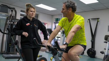 A man in a yellow shirt rides a stationary bike in a gym, smiling and talking with a woman standing beside him. The gym has various exercise equipment and bright lighting.