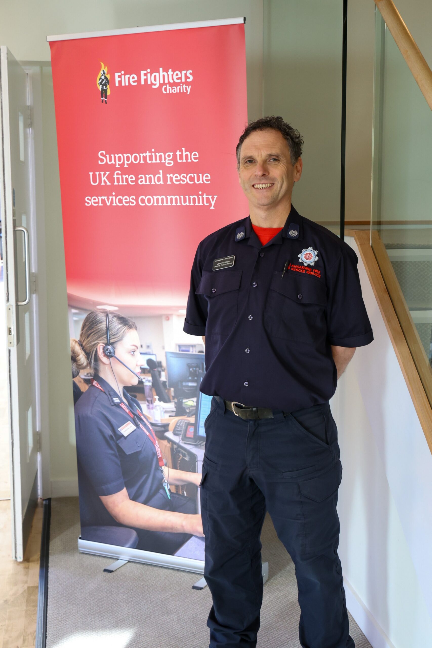 A smiling firefighter in uniform stands indoors next to a Fire Fighters Charity banner that reads, "Supporting the UK fire and rescue services community.