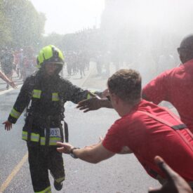 A firefighter in full gear runs through water spray on a sunny street, reaching out to high-five cheering people in red shirts during a public event or race.