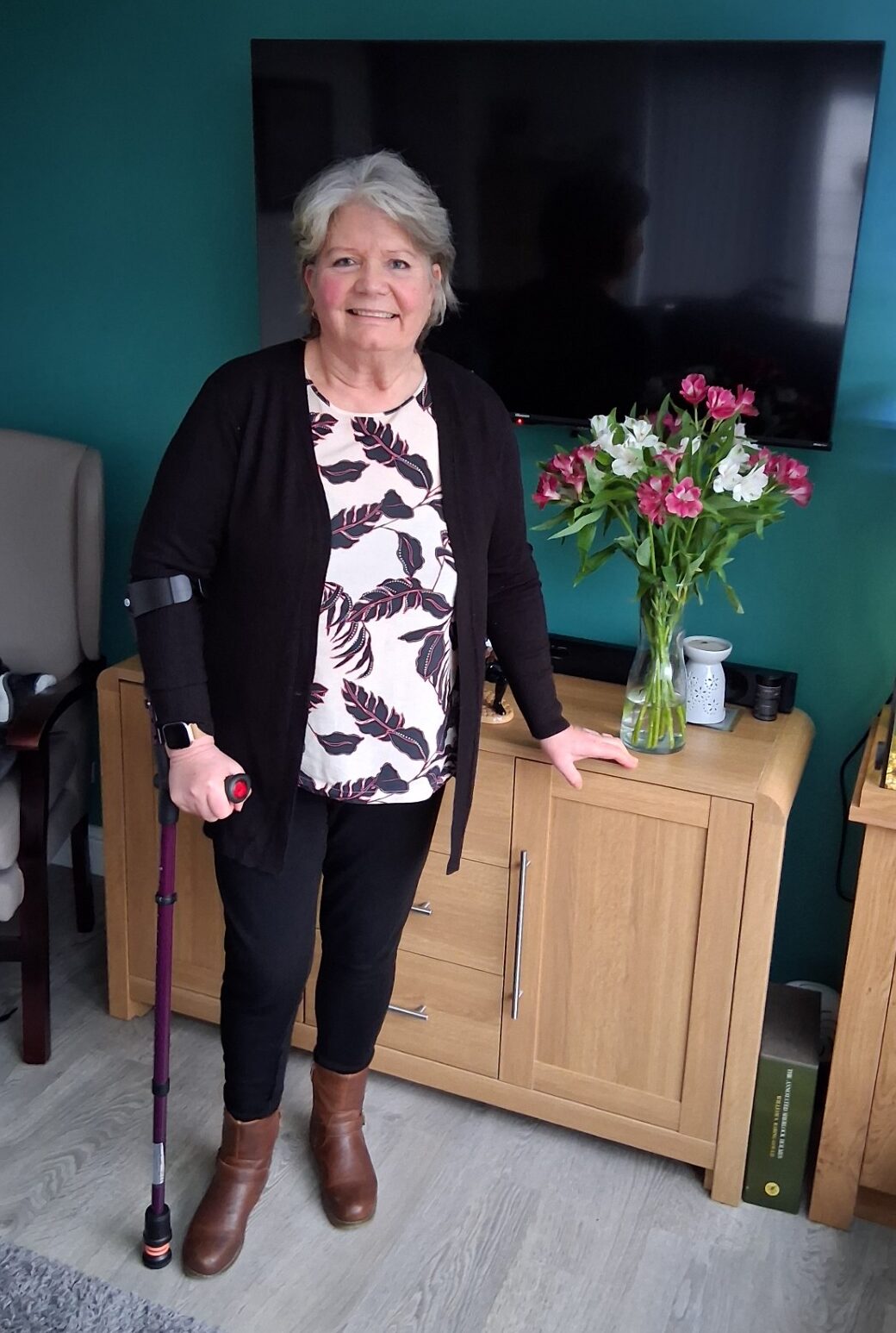 A smiling older woman with gray hair stands indoors, holding a walking stick. She is next to a wooden cabinet with a vase of pink and white flowers and a fish tank, with a large TV mounted on a green wall behind her.