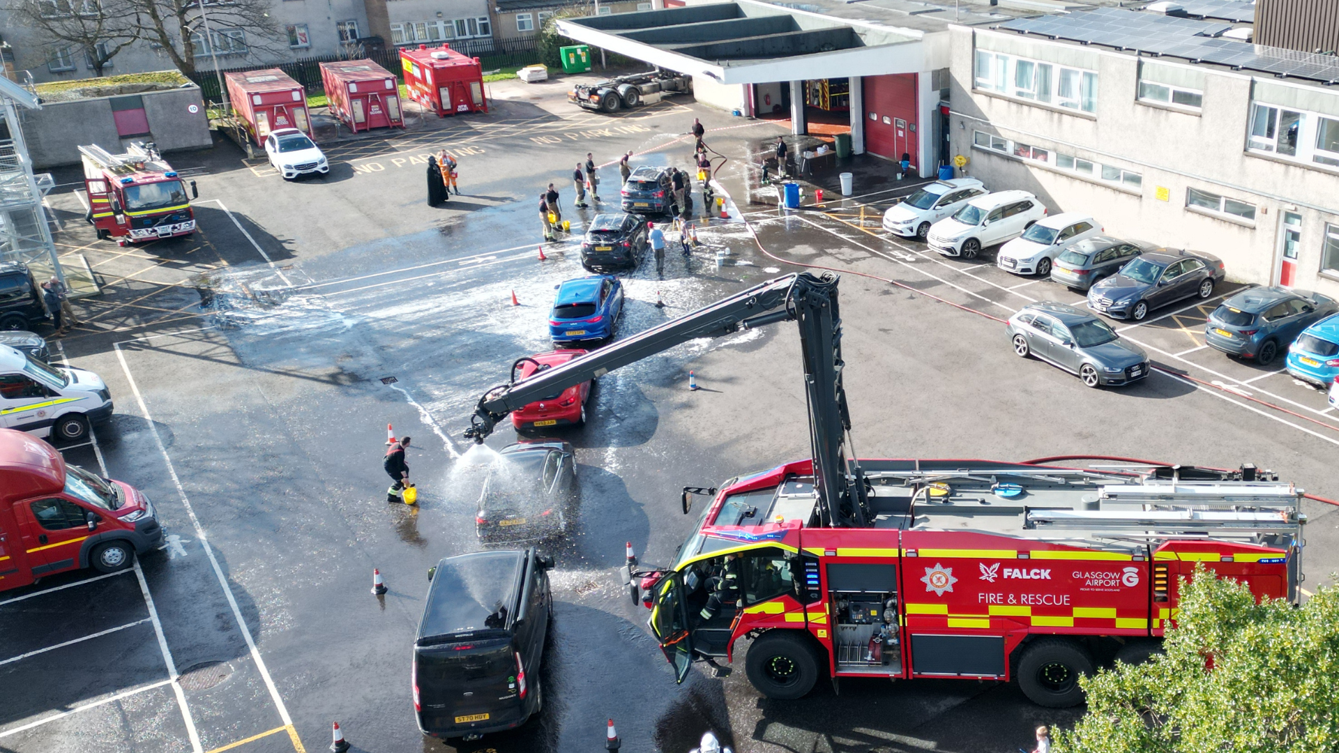 A fire truck sprays water in a parking lot during a training exercise at a fire station, with firefighters and parked cars visible around the area.