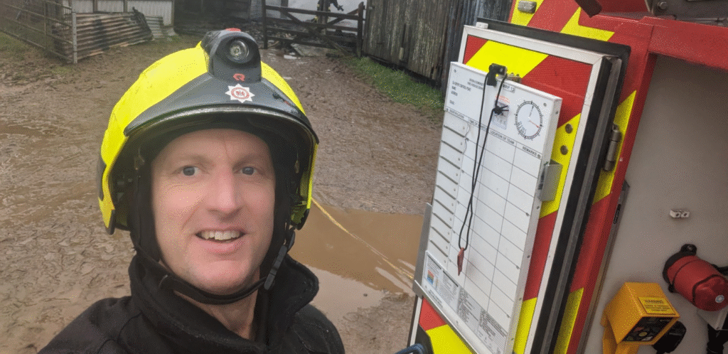A firefighter in a yellow helmet smiles for a selfie outdoors near a muddy area, standing beside an incident command board attached to a fire vehicle.