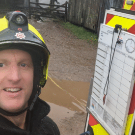 A firefighter in a yellow helmet smiles for a selfie outdoors near a muddy area, standing beside an incident command board attached to a fire vehicle.