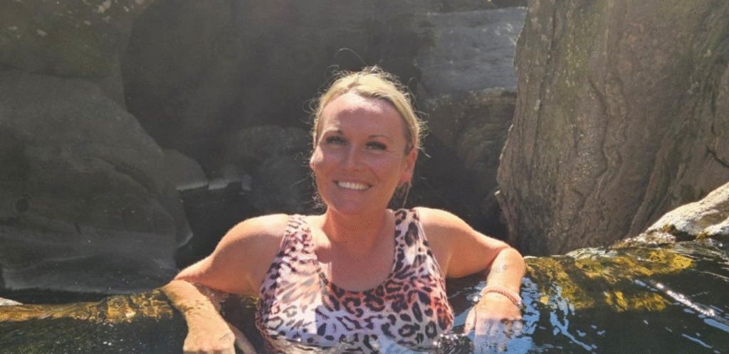 A woman with blond hair in a leopard print swimsuit smiles while relaxing in a natural pool, holding onto rocks with trees and water around her. Sunlight shines on her and the scene.