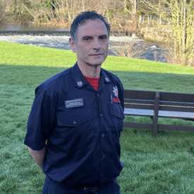 A person in a dark uniform stands on grass near a river, with benches and trees in the background on a sunny day.