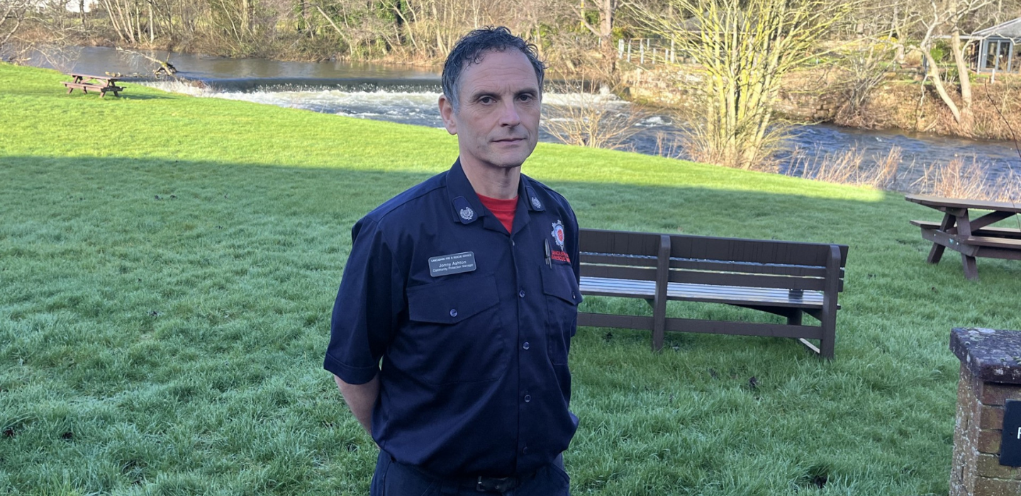 A person in a dark uniform stands on grass near a river, with benches and trees in the background on a sunny day.