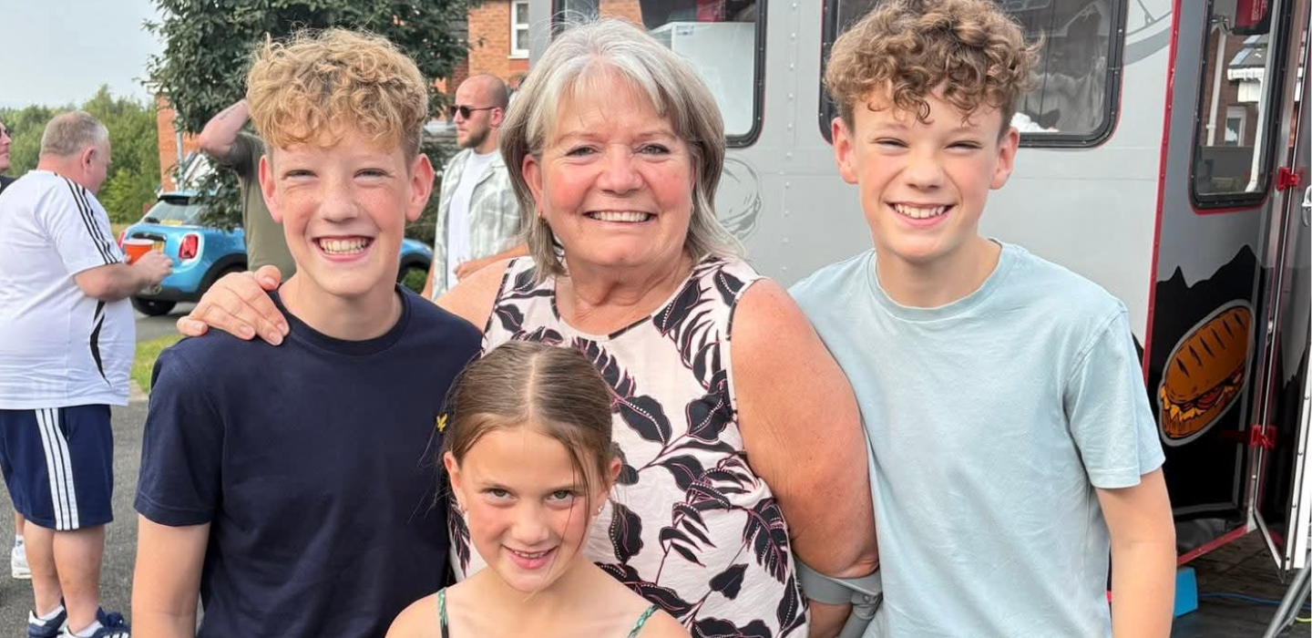 An older woman smiles while standing outdoors with three smiling children—two boys and a girl—on a sunny day, with a food truck and other people visible in the background.