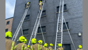 Firefighters in yellow helmets and brown uniforms stand in front of a building, watching colleagues climb tall ladders leading to upper windows during a training exercise.