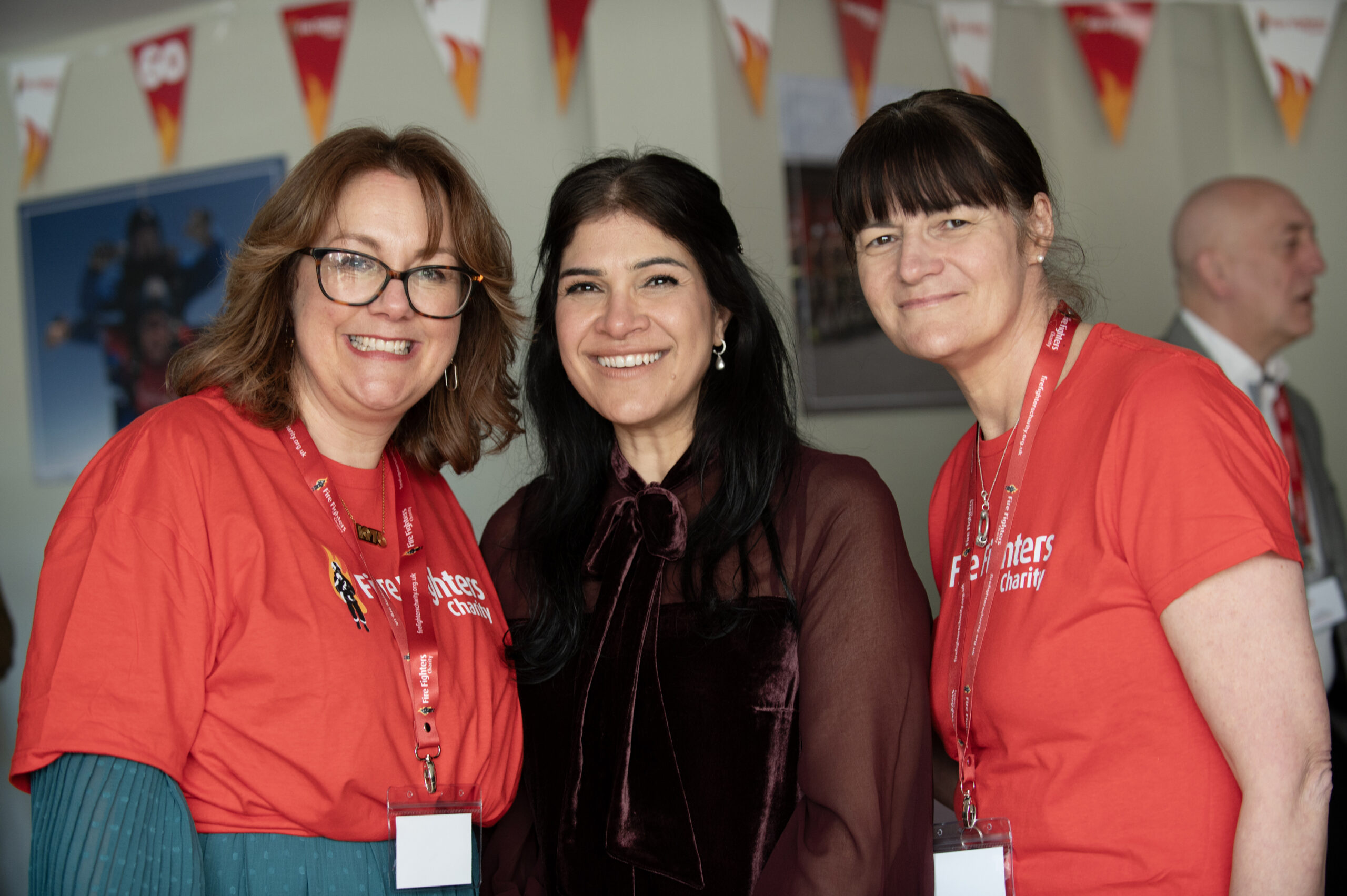 Three women smiling at the camera; two are wearing red shirts with lanyards and name badges, while the third wears a dark blouse. There are colorful triangular banners hanging in the background.