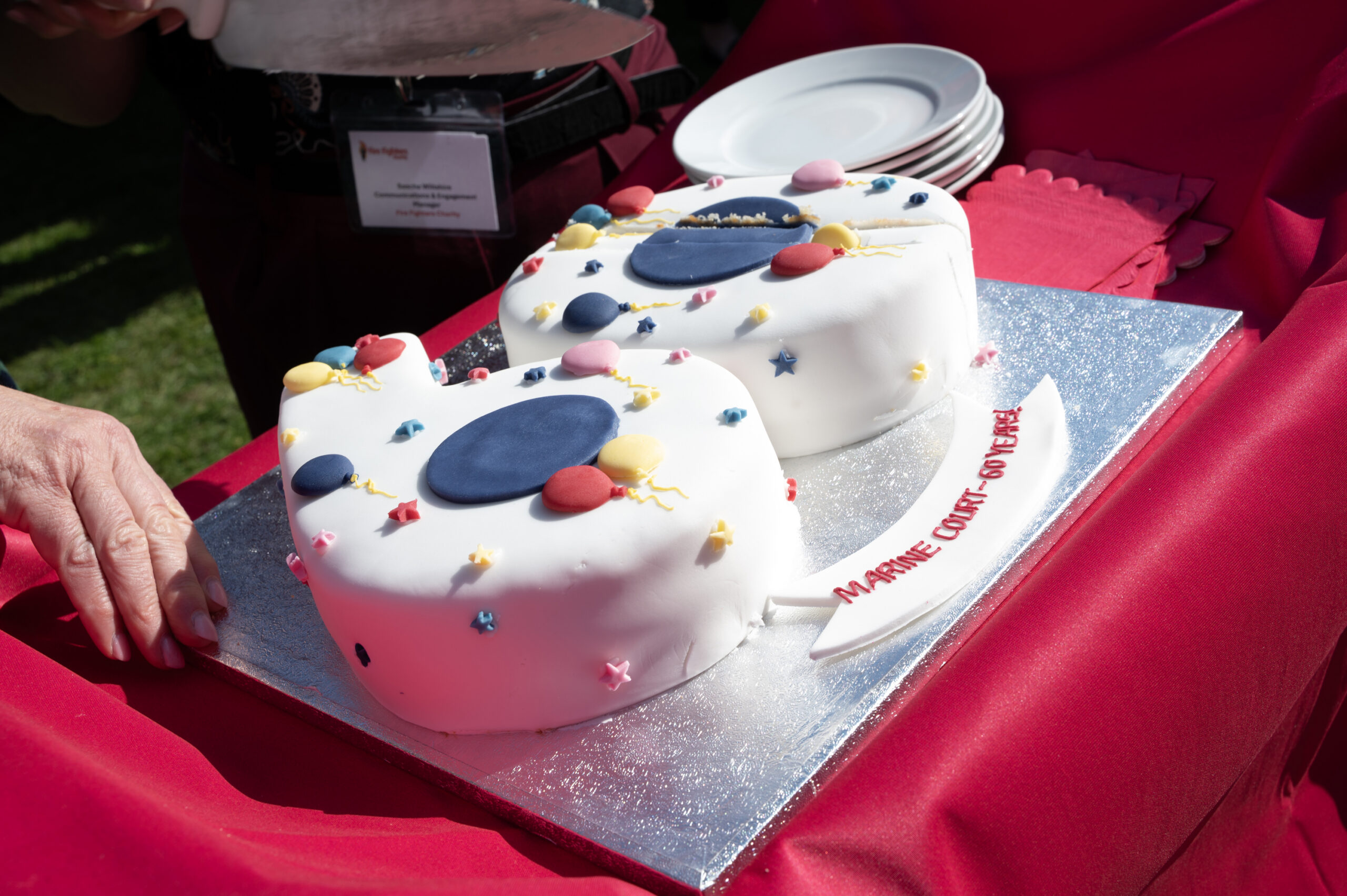 A heart-shaped cake decorated with colorful stars and circles sits on a silver board with a label reading "MARINE COURT 50 YEARS." Plates and a serving utensil are nearby on a red tablecloth.
