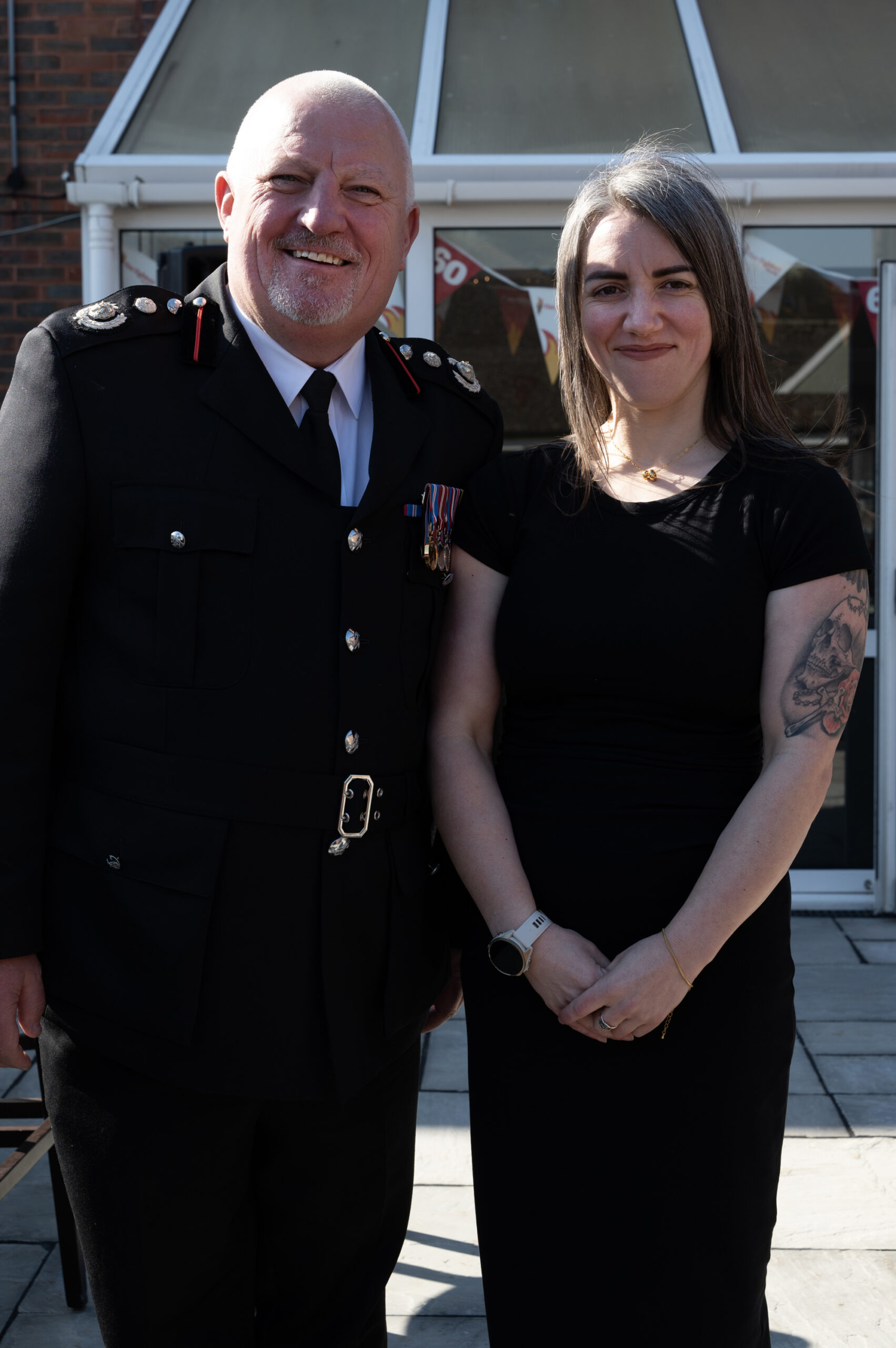 A man in a formal dark uniform with medals stands smiling next to a woman in a black dress with a tattoo on her arm. They are outdoors in front of a building with large glass windows.