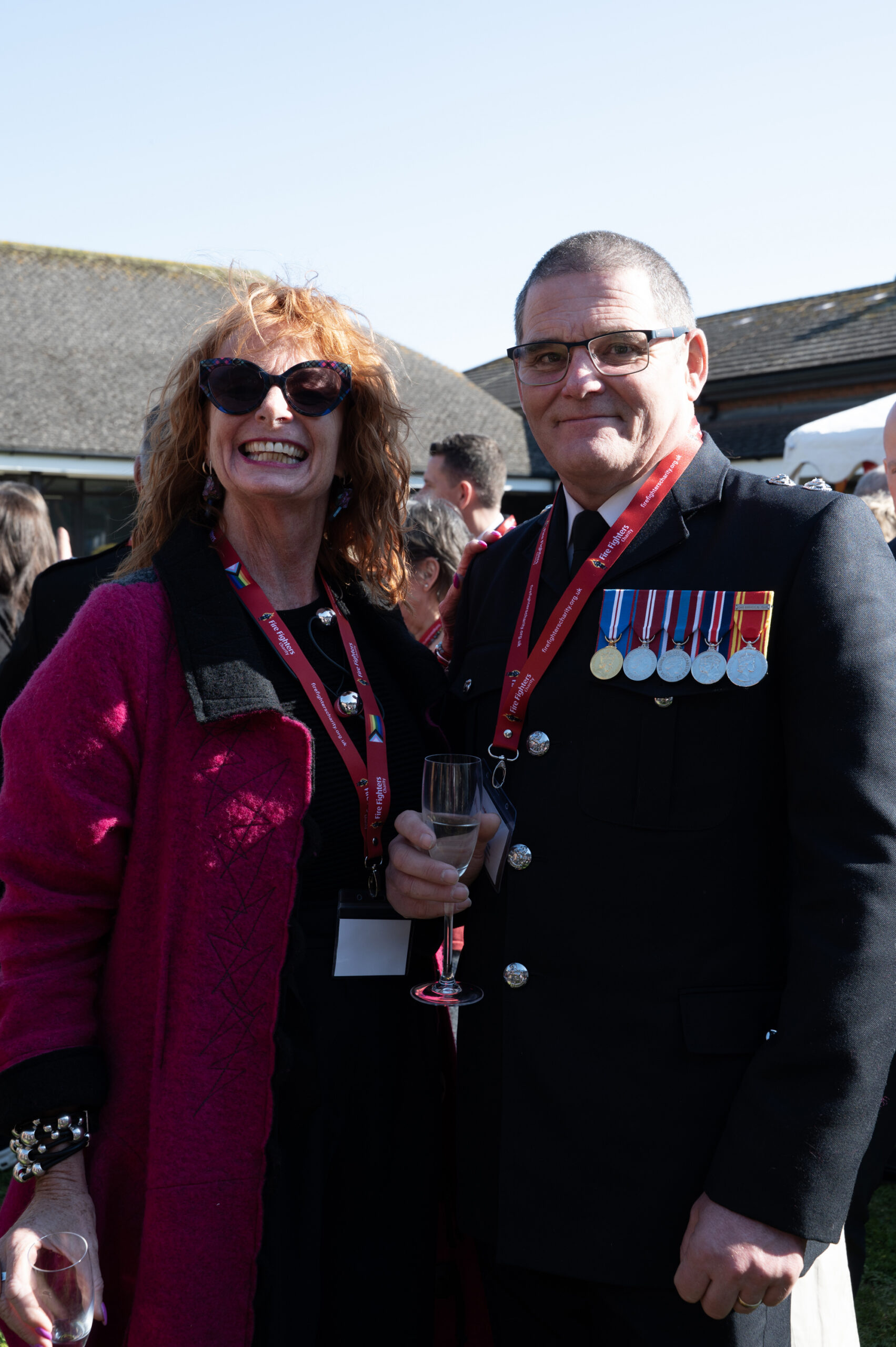 A smiling woman in sunglasses and a red coat stands next to a man in a decorated uniform with medals, both holding drinks, at an outdoor event with other people in the background.