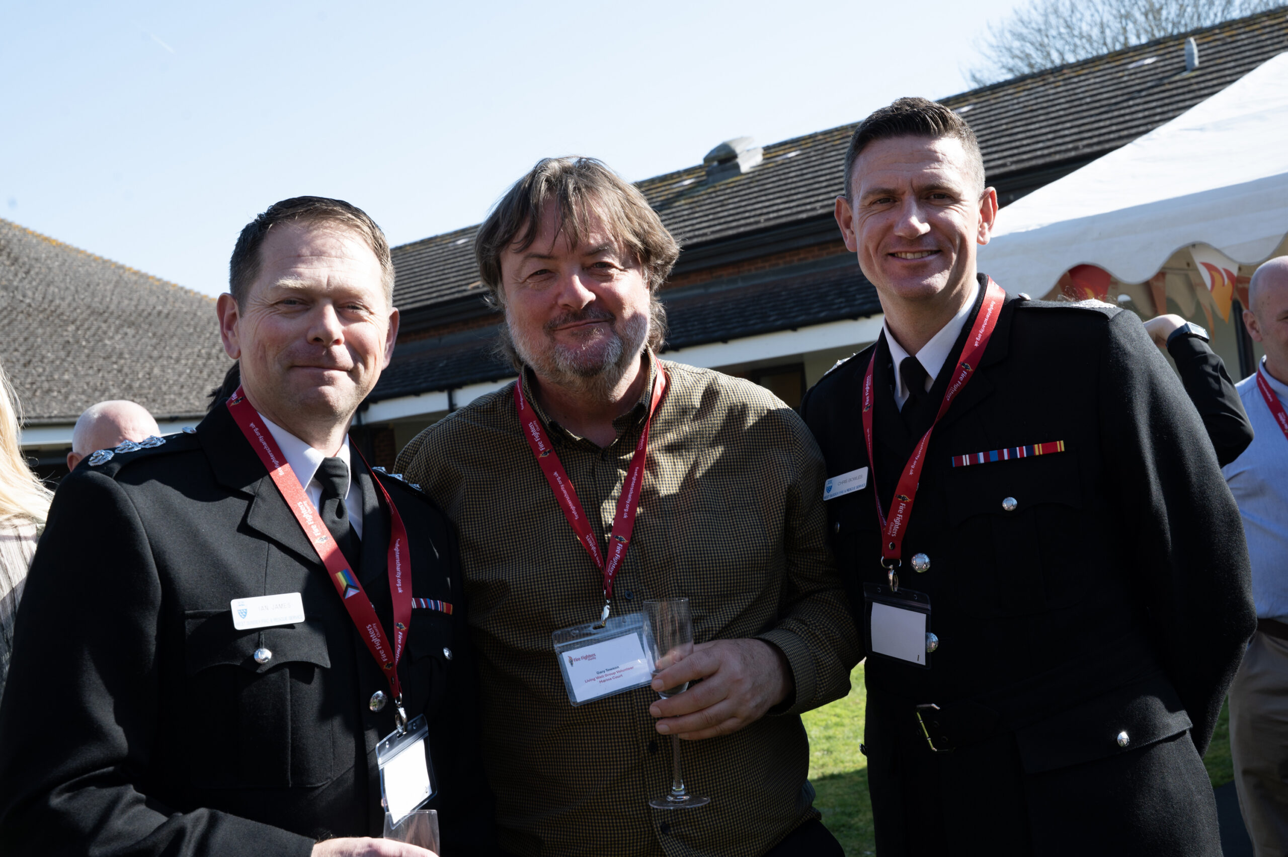 Three men pose outdoors at a daytime event. Two wear black formal uniforms with medals and red lanyards; the man in the center wears a brown shirt, red lanyard, and holds a glass. People and tents are visible in the background.