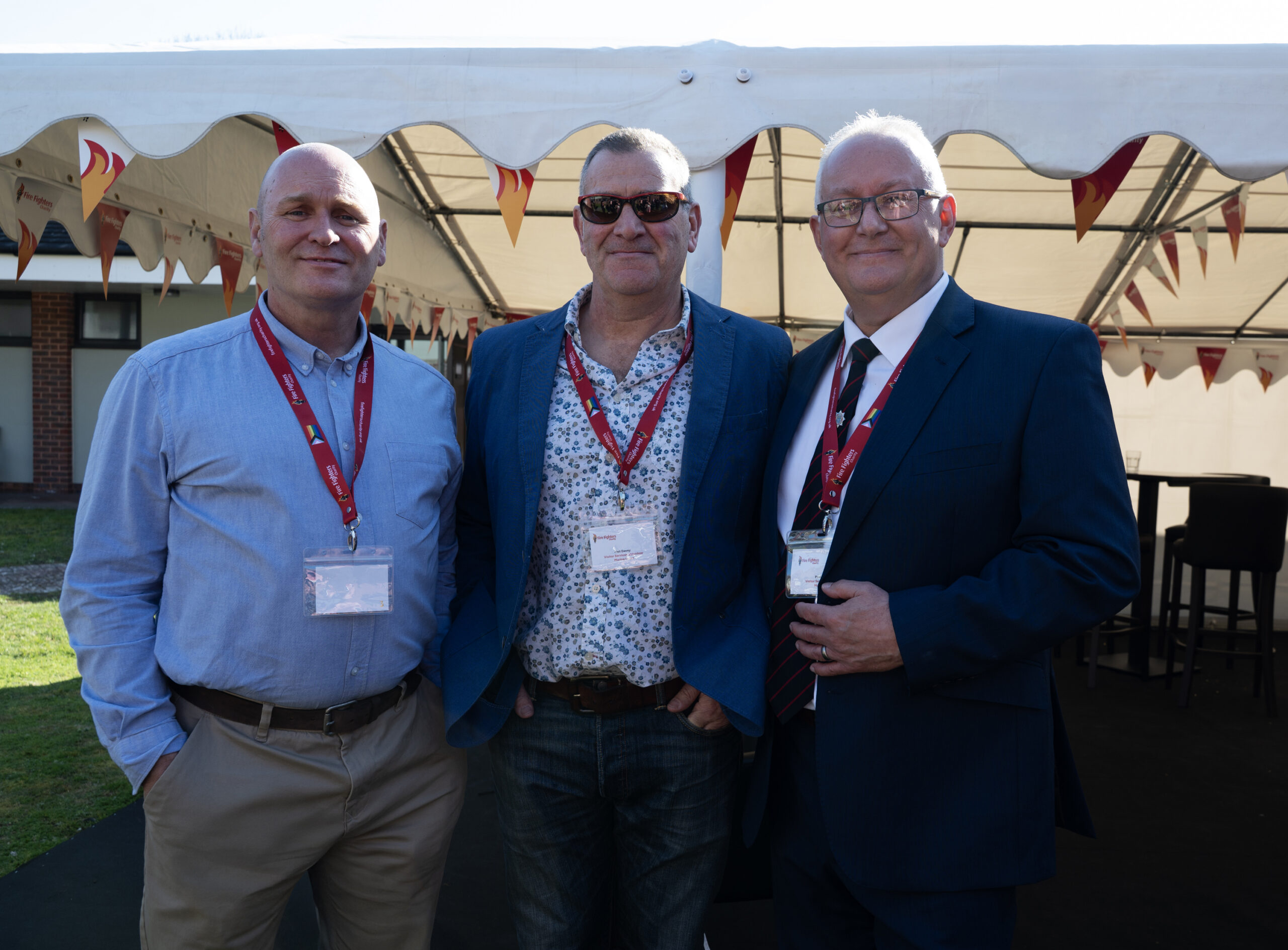 Three men wearing name badges and red lanyards stand together under a white outdoor tent. Two are in business attire, one wears a casual shirt. They are smiling and posing for the camera at an event.