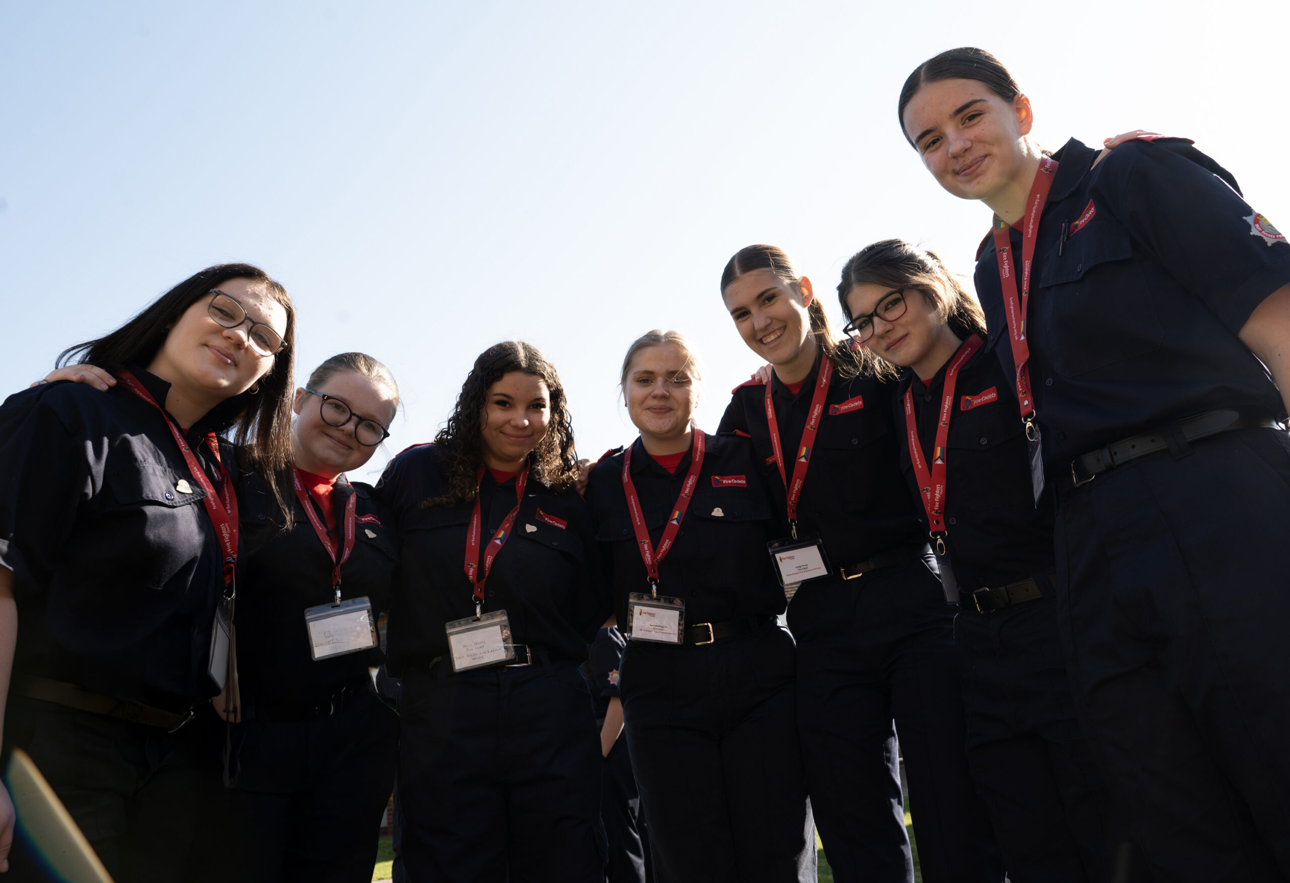 A group of seven young women in matching dark uniforms with red lanyards and name badges stand together outdoors, smiling at the camera under a bright sky.