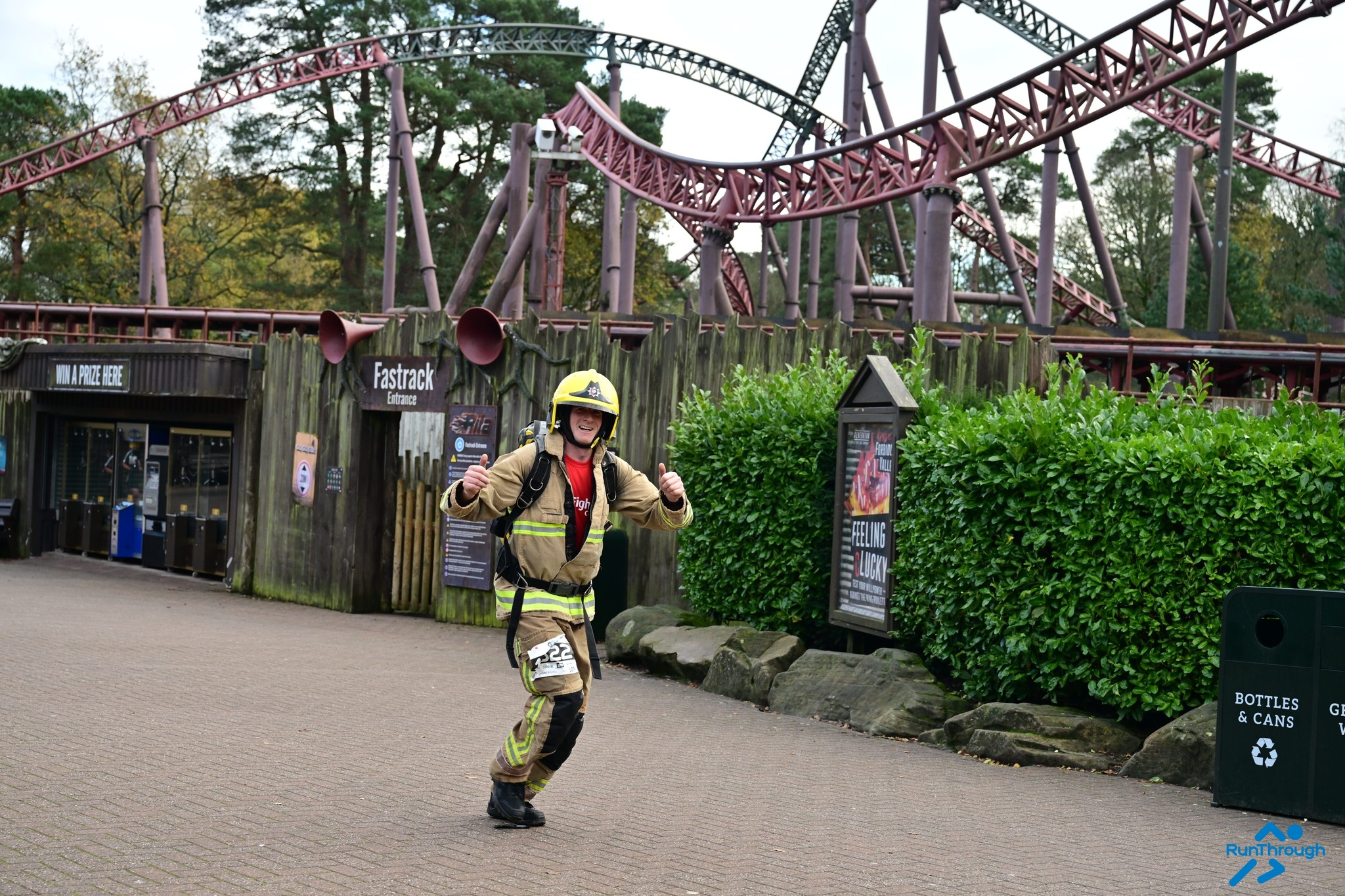 A person in firefighter gear smiles and gives two thumbs up while running in front of a roller coaster at an amusement park. Greenery and a wooden fence are in the background.