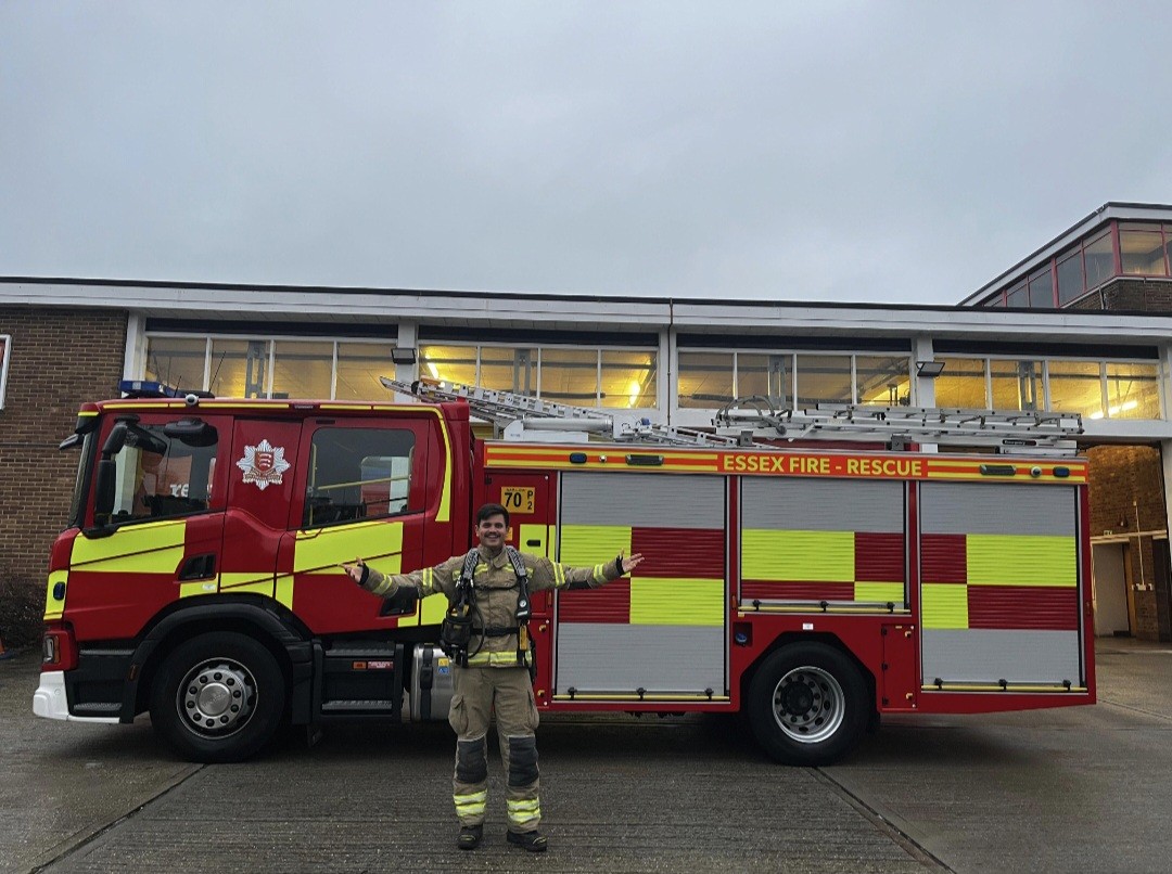 A firefighter in full gear stands smiling with arms outstretched in front of a red and yellow Essex Fire and Rescue truck parked outside a brick fire station.