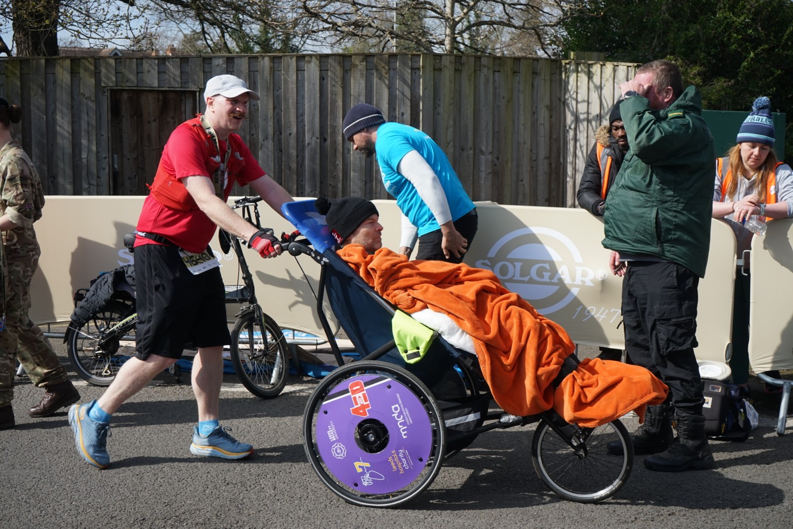 A man in a red shirt pushes a person in an adaptive wheelchair, covered by an orange blanket, during a race. Several people, some cheering and some in athletic gear, stand and walk nearby.