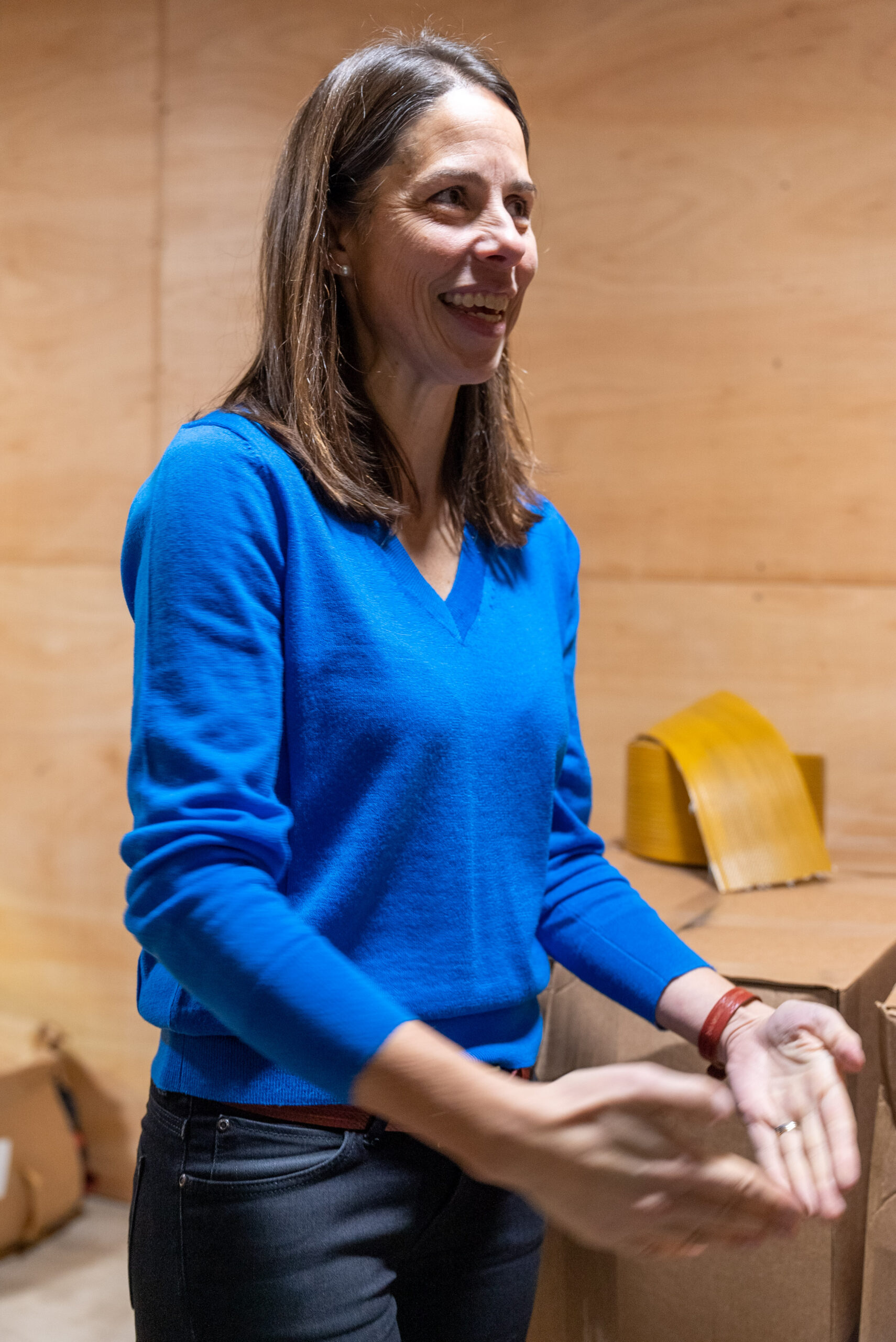 A woman with long brown hair, wearing a bright blue sweater and black pants, stands indoors near cardboard boxes, smiling and gesturing with her hands. The background is wooden paneling.