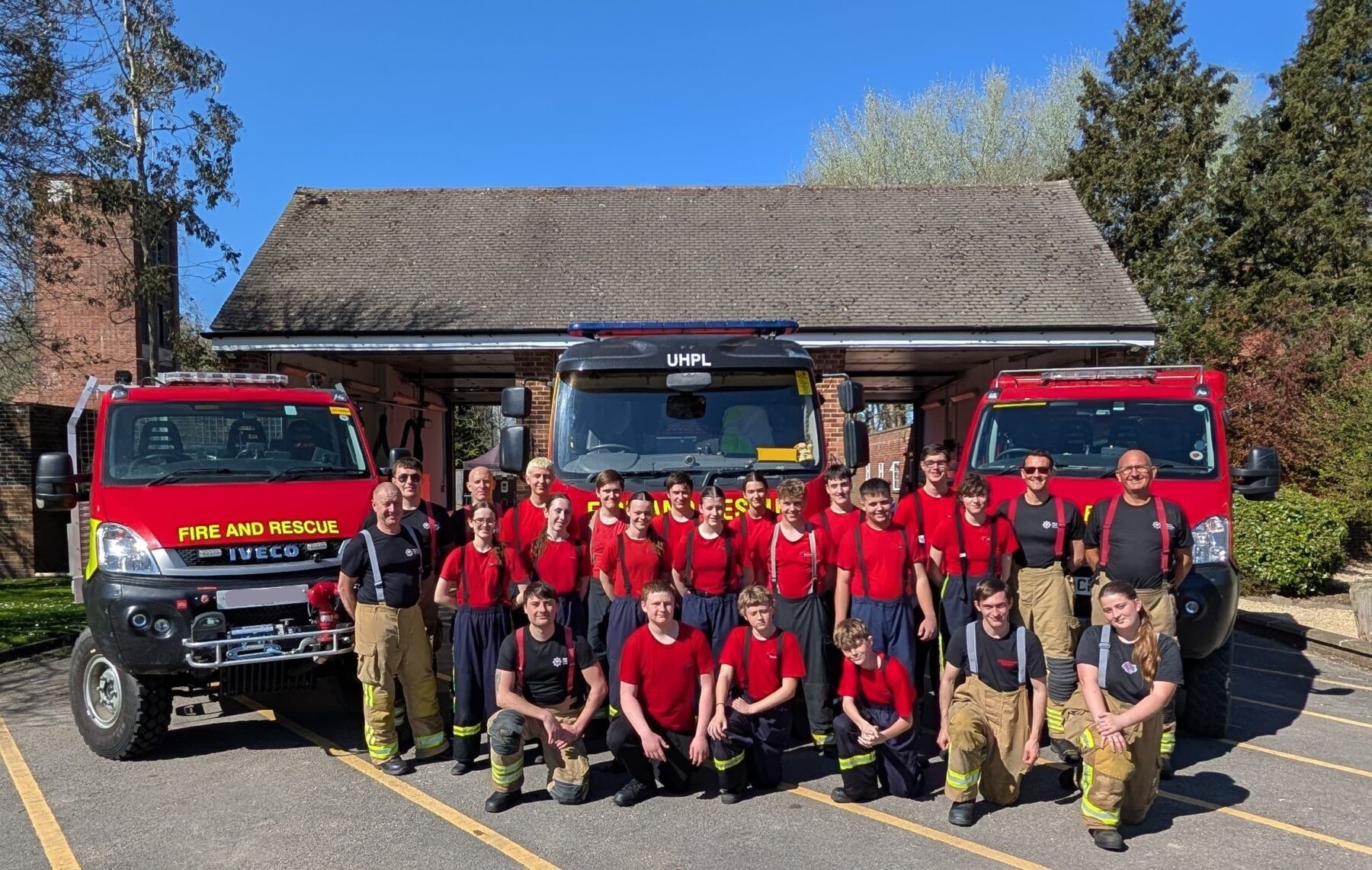 A group of firefighters in red shirts and yellow pants pose in front of three Fire and Rescue trucks, smiling outdoors on a sunny day, with trees and a building in the background.
