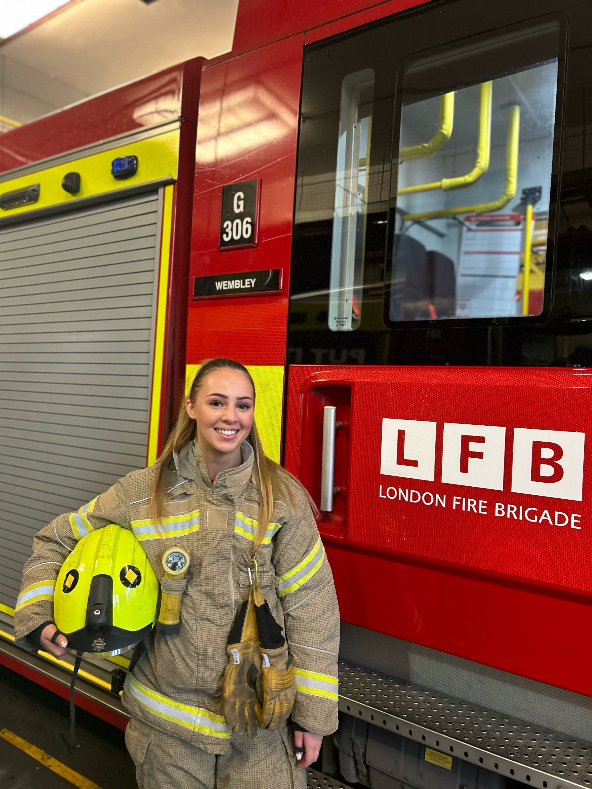 A firefighter in uniform smiles while holding a yellow helmet, standing in front of a red London Fire Brigade fire engine at a station.