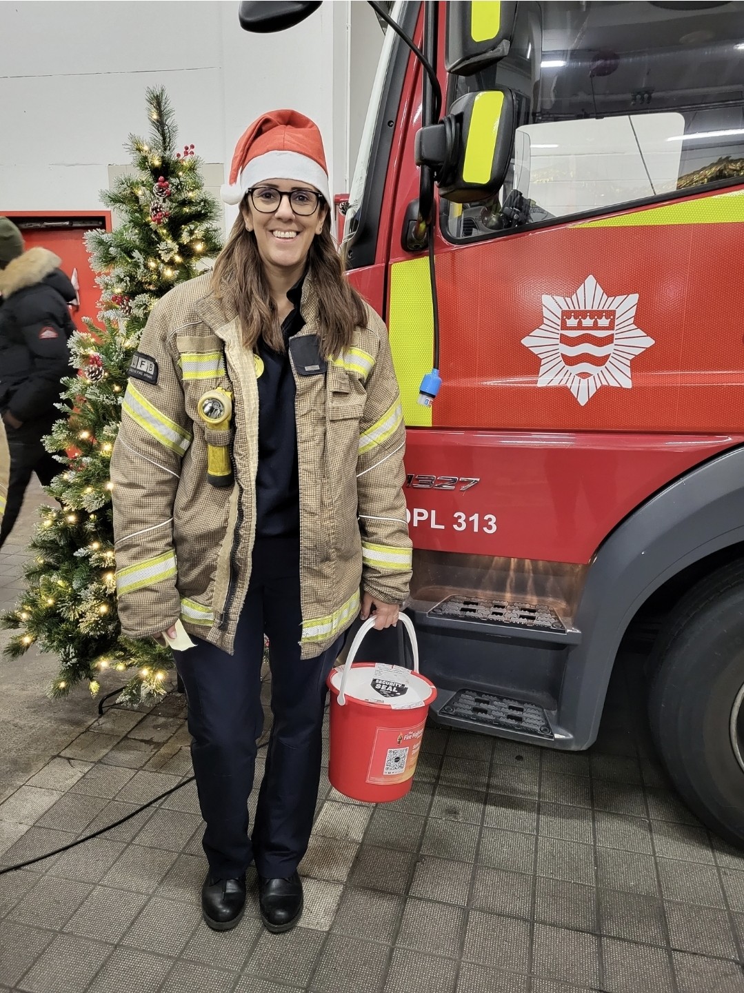A smiling firefighter wearing a Santa hat stands in front of a fire truck, holding a red donation bucket. A decorated Christmas tree is visible beside her.