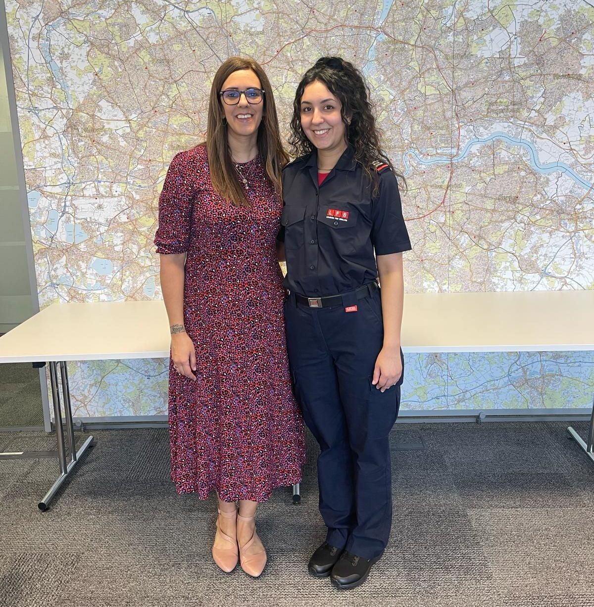 Two women stand side by side in front of a large map. One wears a patterned dress and glasses, while the other wears a dark firefighter uniform. Both are smiling and looking at the camera.