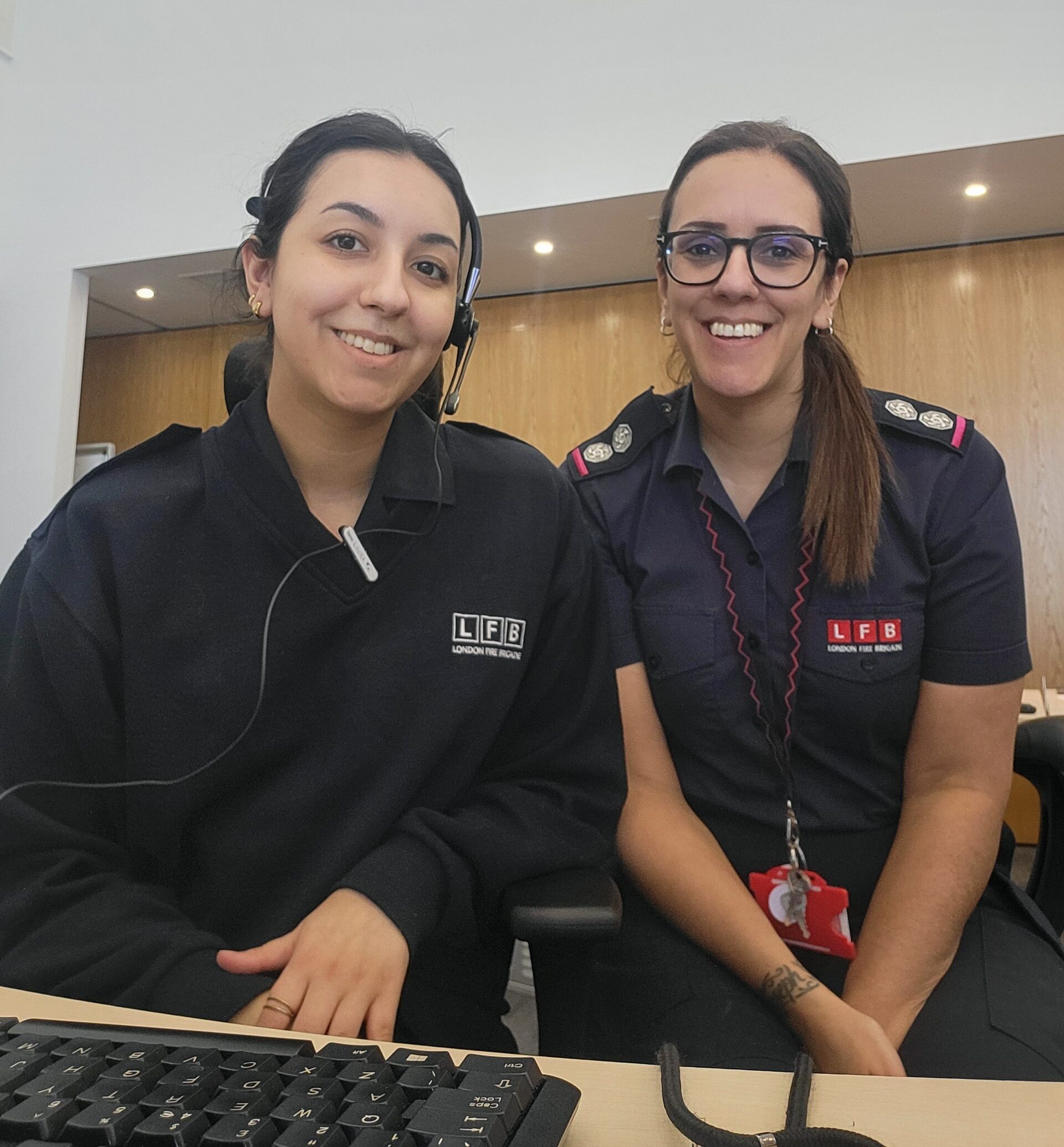 Two women wearing LFB uniforms sit at a desk in an office, smiling at the camera. One woman has a headset on, and a computer keyboard is visible in the foreground.