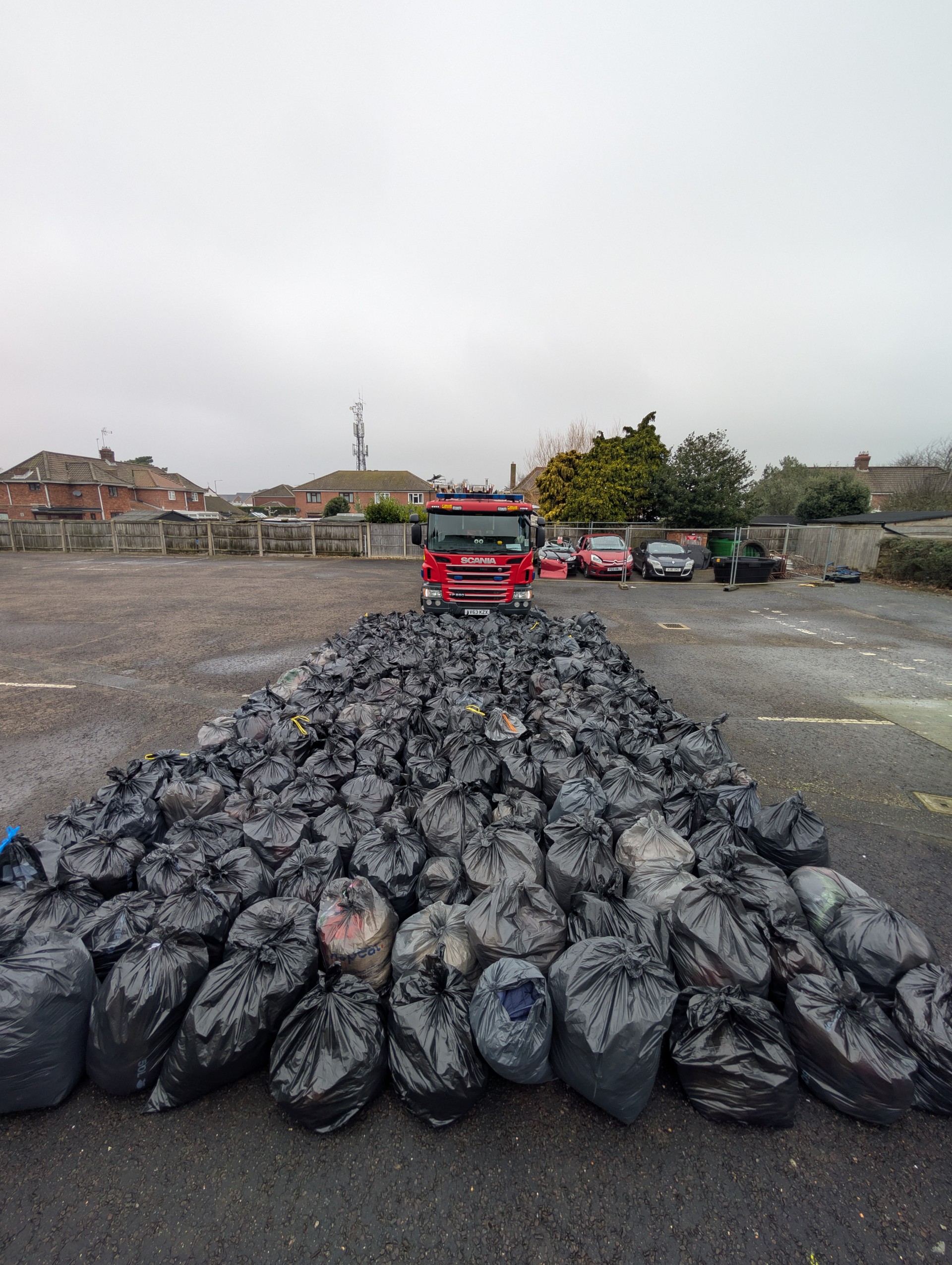 A red garbage truck is parked in a lot, facing rows of numerous black trash bags neatly lined up on the ground, with houses, cars, and a cloudy sky in the background.