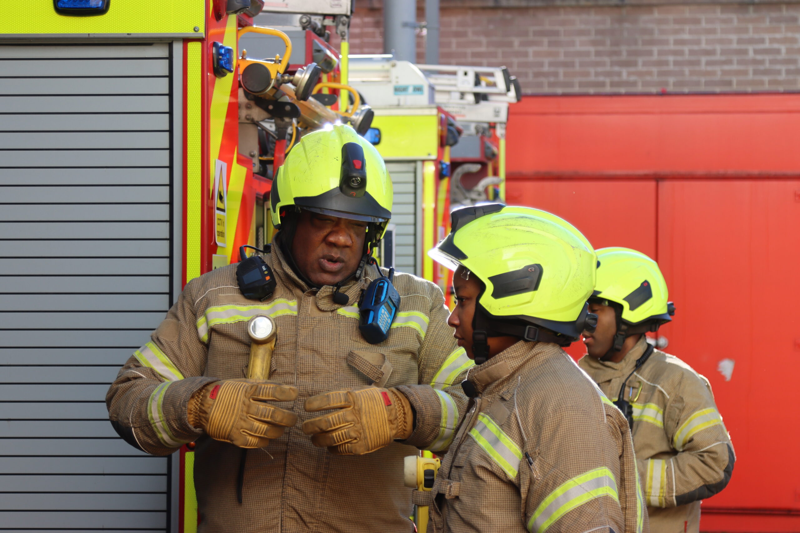 Three firefighters in yellow helmets and brown protective gear stand beside a fire truck, engaged in conversation. Two are facing each other, while a third is in the background, all near red and grey fire station doors.