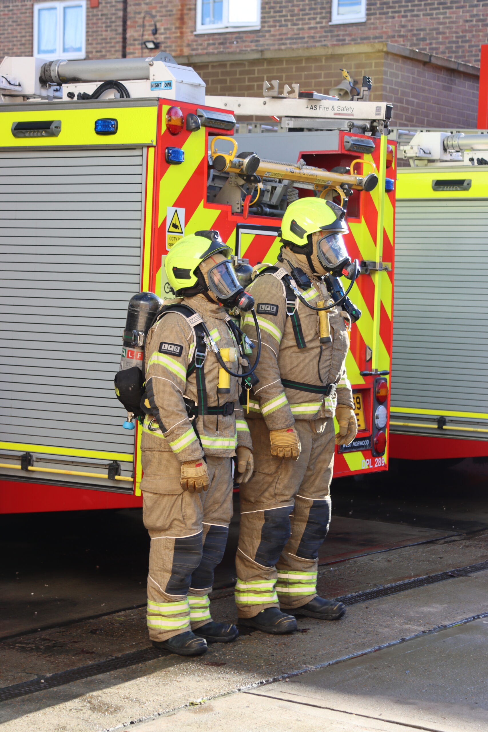 Two firefighters in full gear, including helmets and breathing apparatus, stand side by side next to a fire truck with red and yellow reflective markings, outside a building on a sunny day.