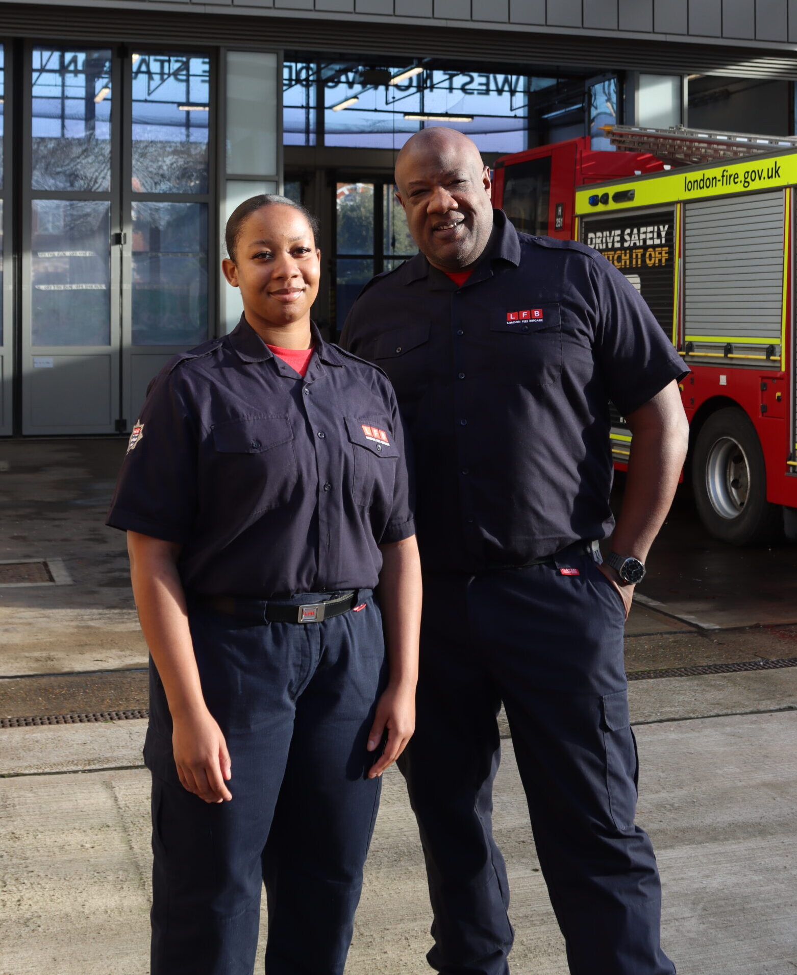 Two firefighters in uniform stand outside a fire station, with a red fire engine visible in the background. They are posing and smiling for the camera on a sunny day.