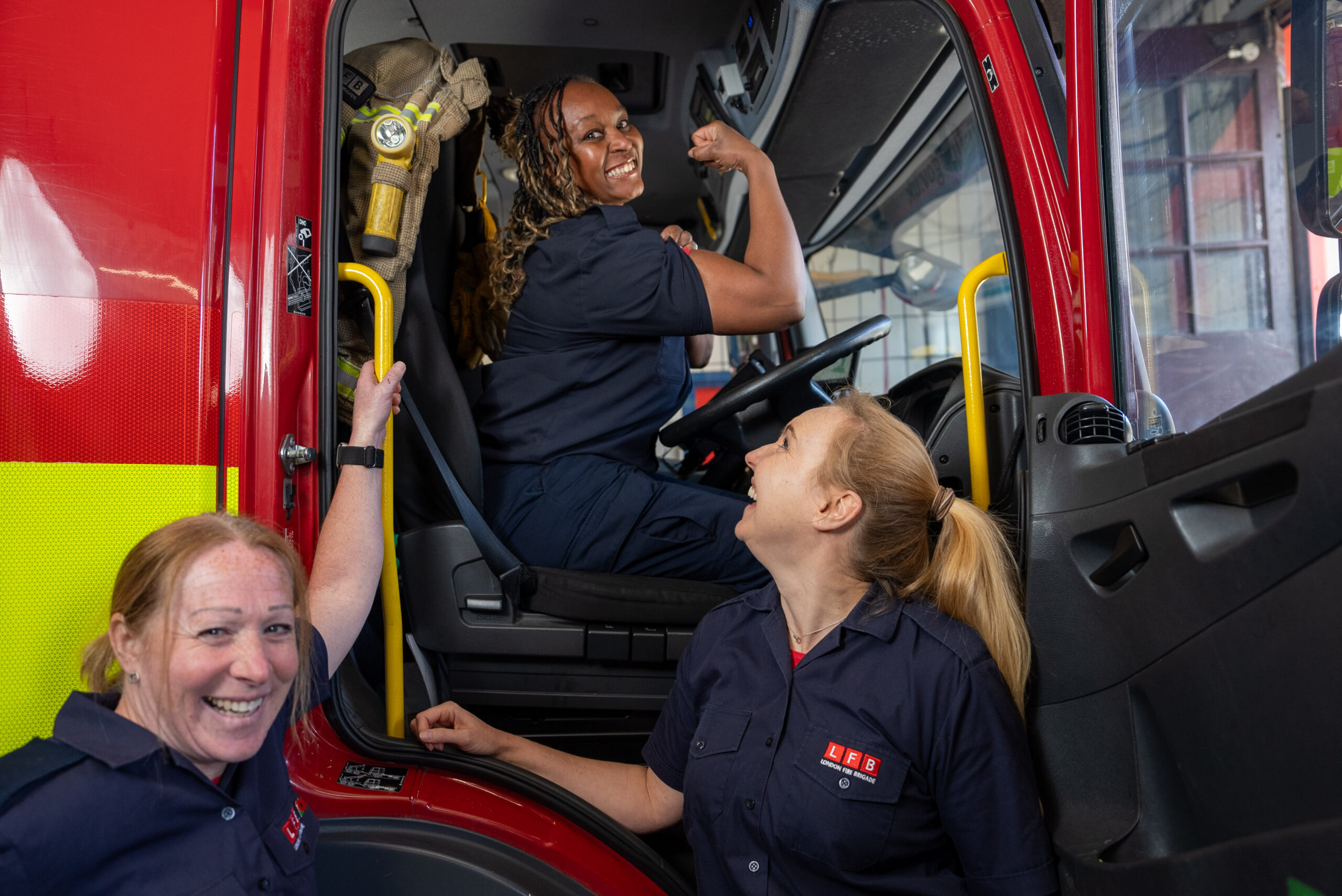 Three female firefighters in uniform pose cheerfully inside and beside a fire truck. One woman sits in the driver's seat flexing her arm, while the other two smile and look up at her, highlighting teamwork and camaraderie.