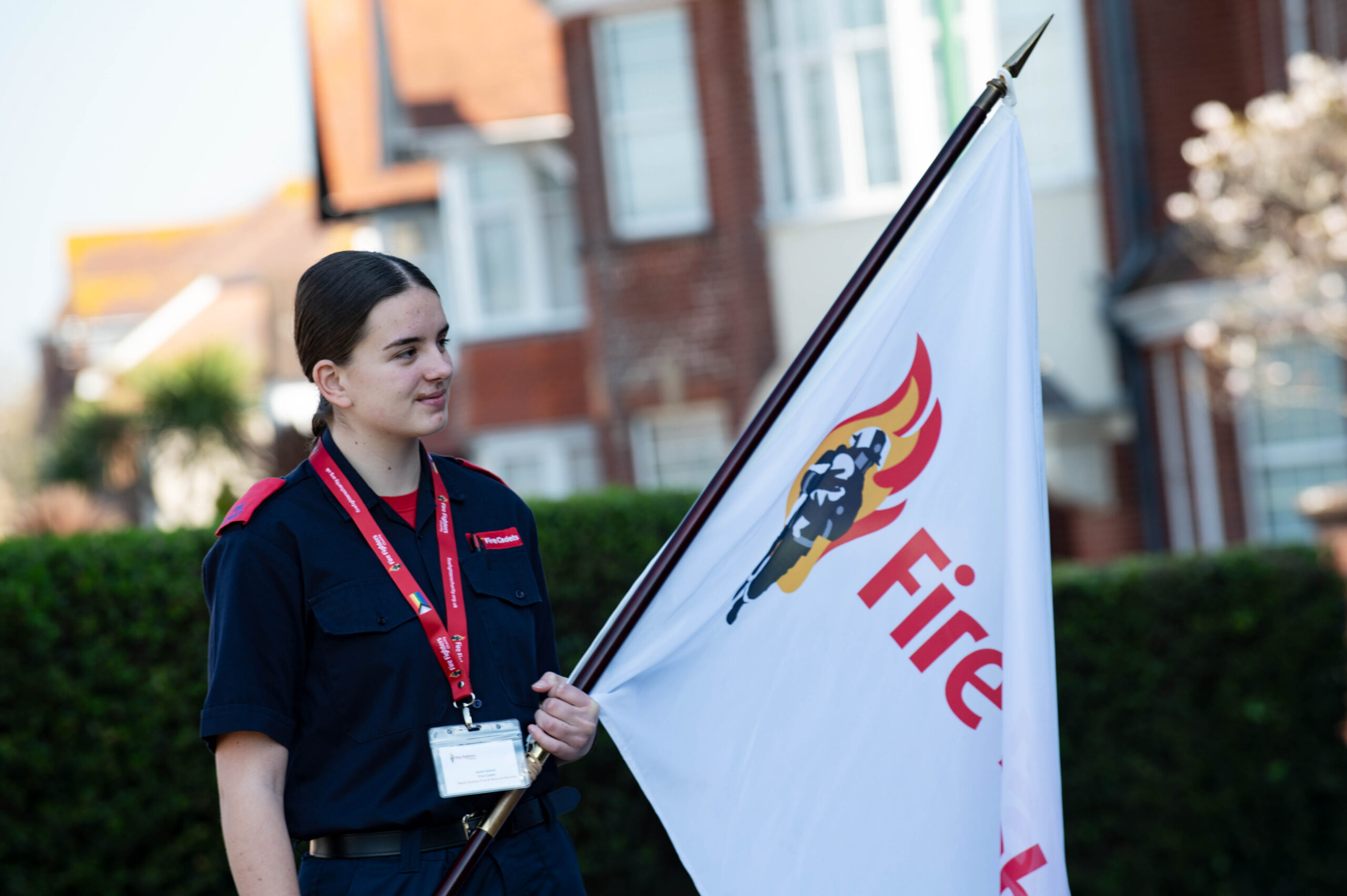 A young person in a navy blue uniform holds a white flag with a fire service logo and the word "Fire" on it, standing outdoors in front of houses and greenery.
