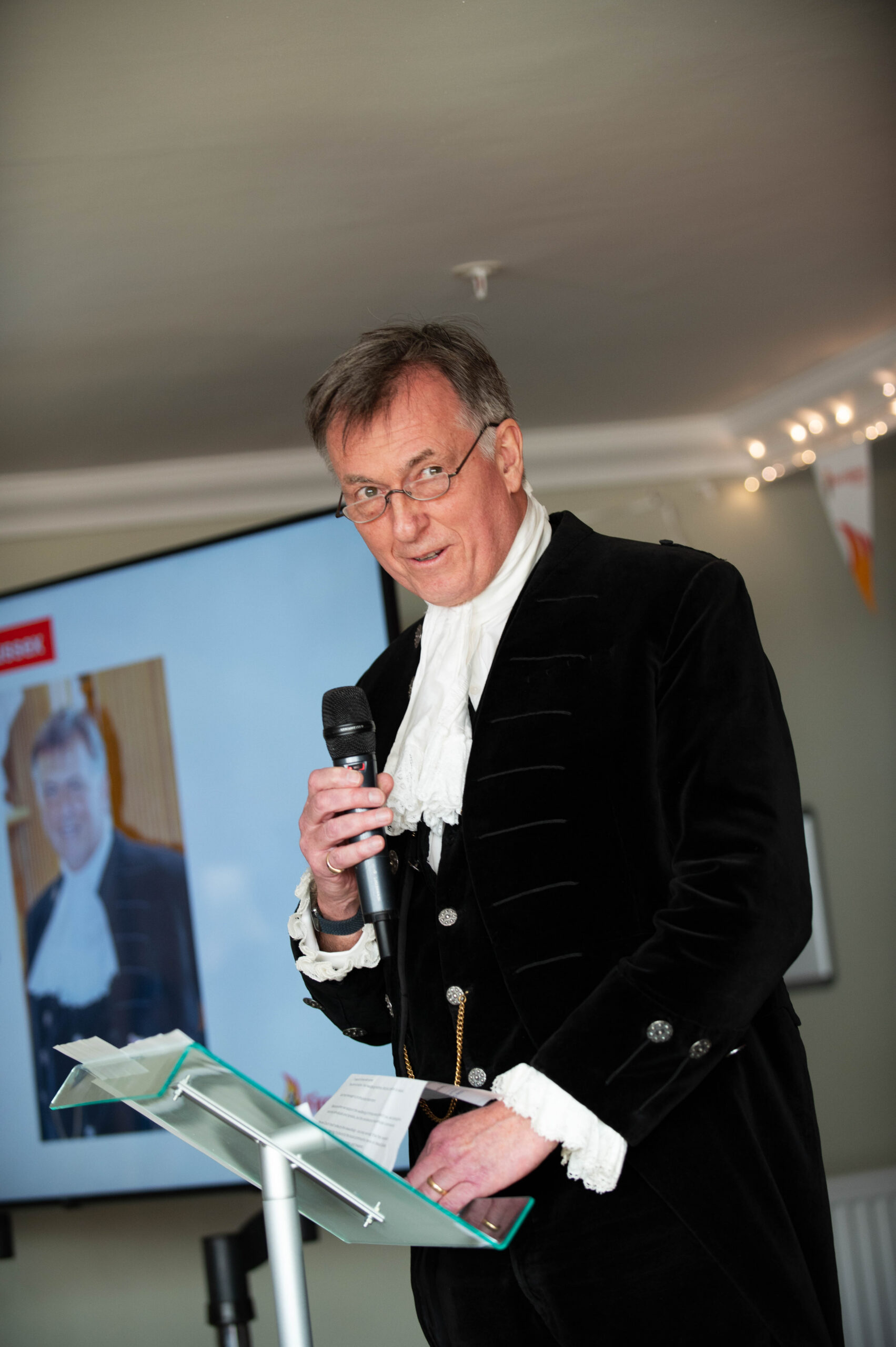 A man in formal, historical clothing speaks into a microphone while standing at a podium. A screen behind him displays a photo of the same man in similar attire. The setting appears to be indoors with soft lighting.