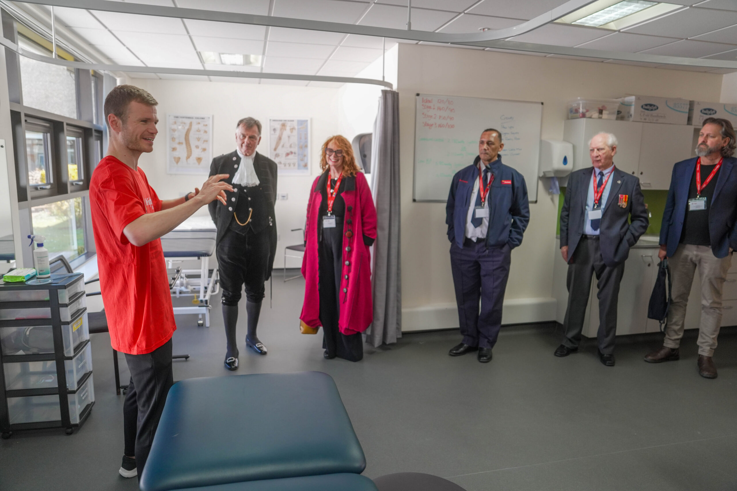 A man in a red shirt gestures while speaking to five adults standing in a medical examination room; some wear formal attire and name badges, and anatomy charts are visible on the wall behind them.