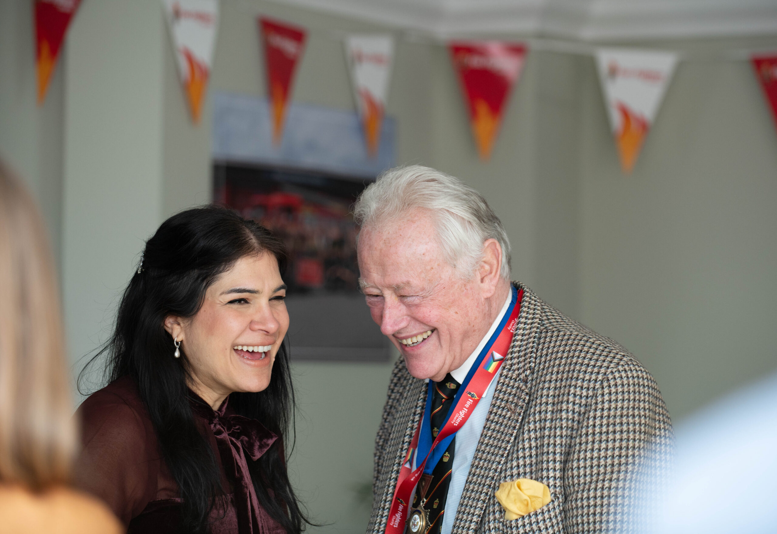 A woman and an older man, both smiling and laughing, stand together indoors. The man wears medals around his neck and a checked jacket with a yellow pocket square. Red and white bunting is visible in the background.