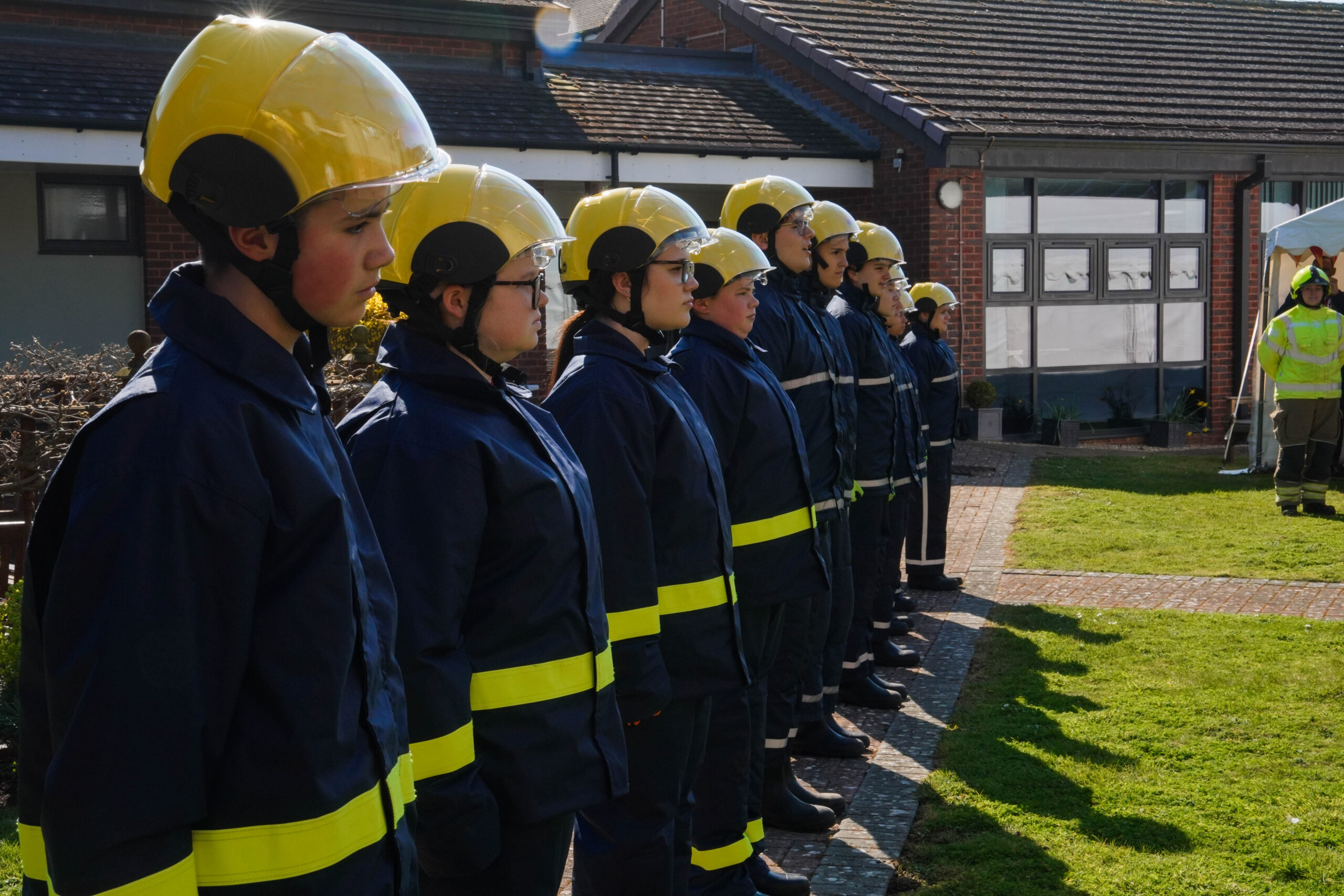 A row of firefighters in yellow helmets and dark uniforms stand in formation outdoors, facing a person in safety gear near a building on a sunny day.