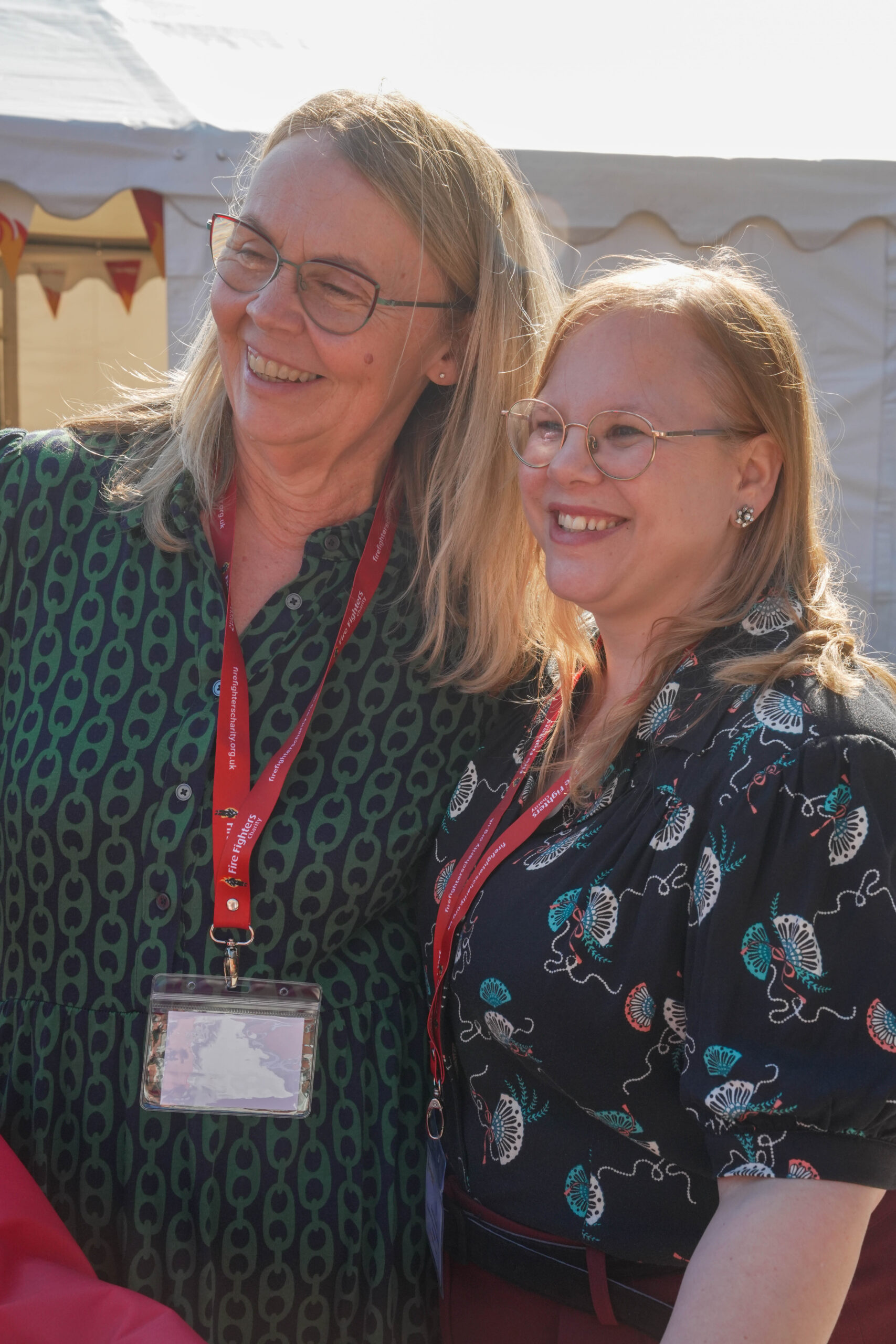 Two smiling women with glasses stand close together outdoors, both wearing patterned shirts and red lanyards with name badges, in front of a white event tent.