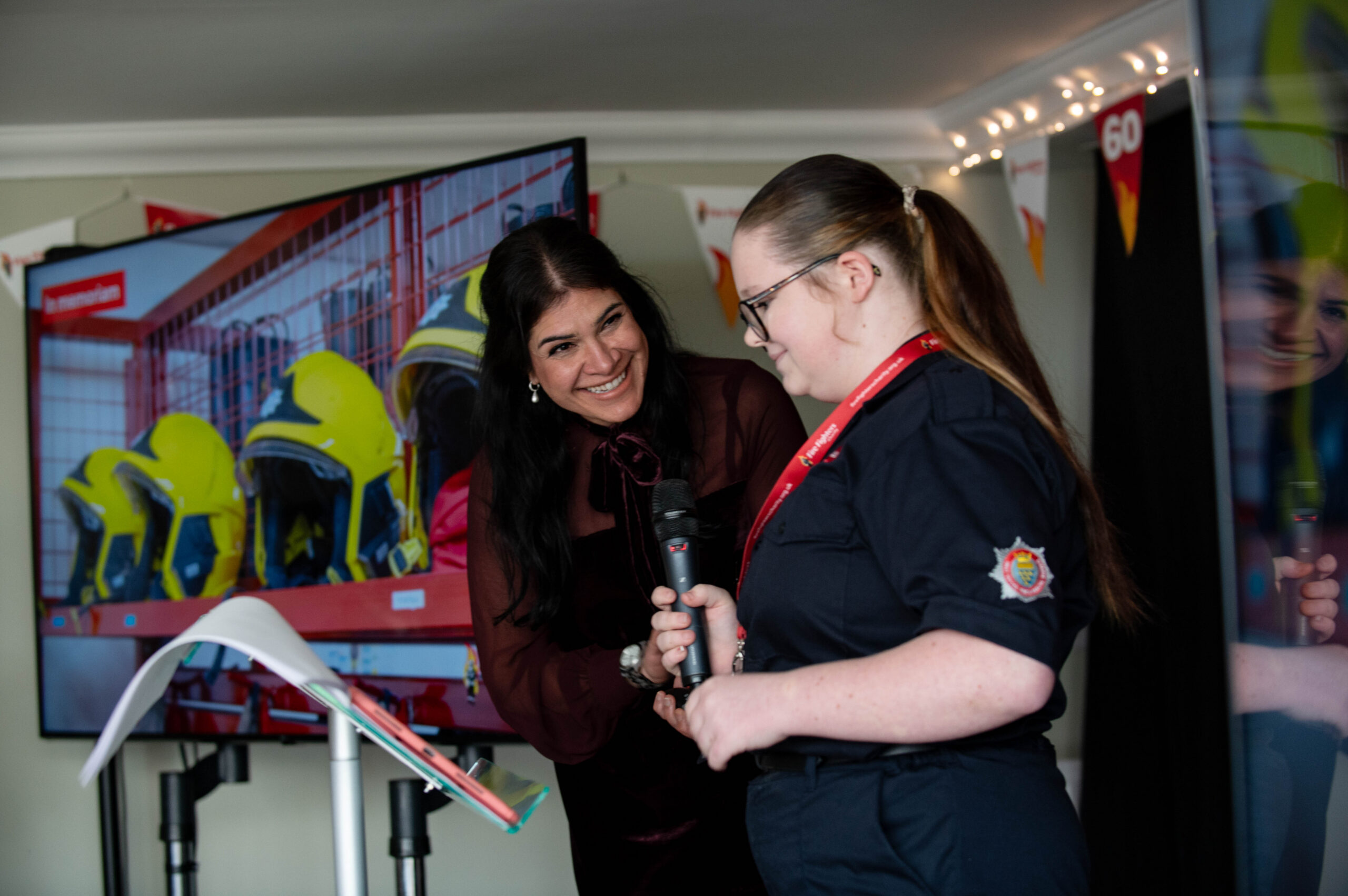 A woman smiles at a young girl in a fire service uniform who holds a microphone. They stand near a podium, with a screen showing yellow firefighter helmets in the background.