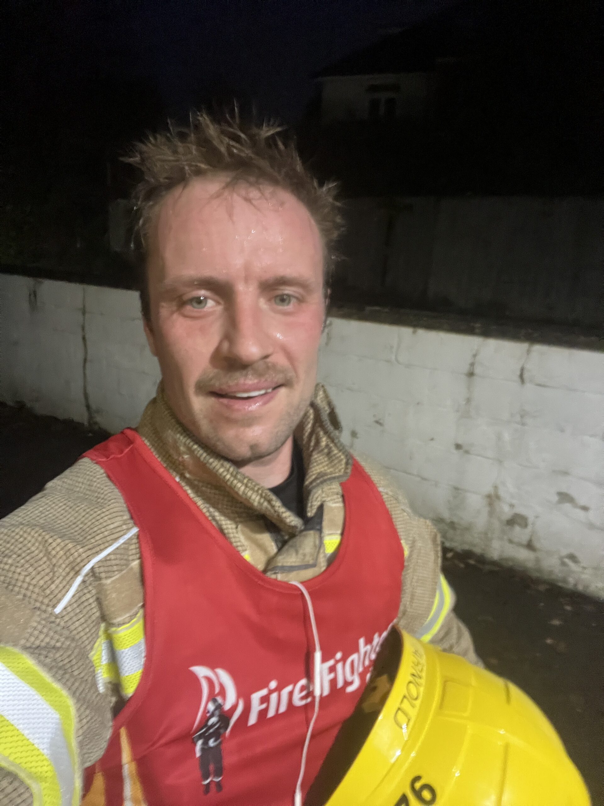 A firefighter in uniform and a red vest, holding a yellow helmet, smiles at the camera outdoors at night. He appears sweaty, suggesting recent physical activity. A white wall and dark background are visible behind him.