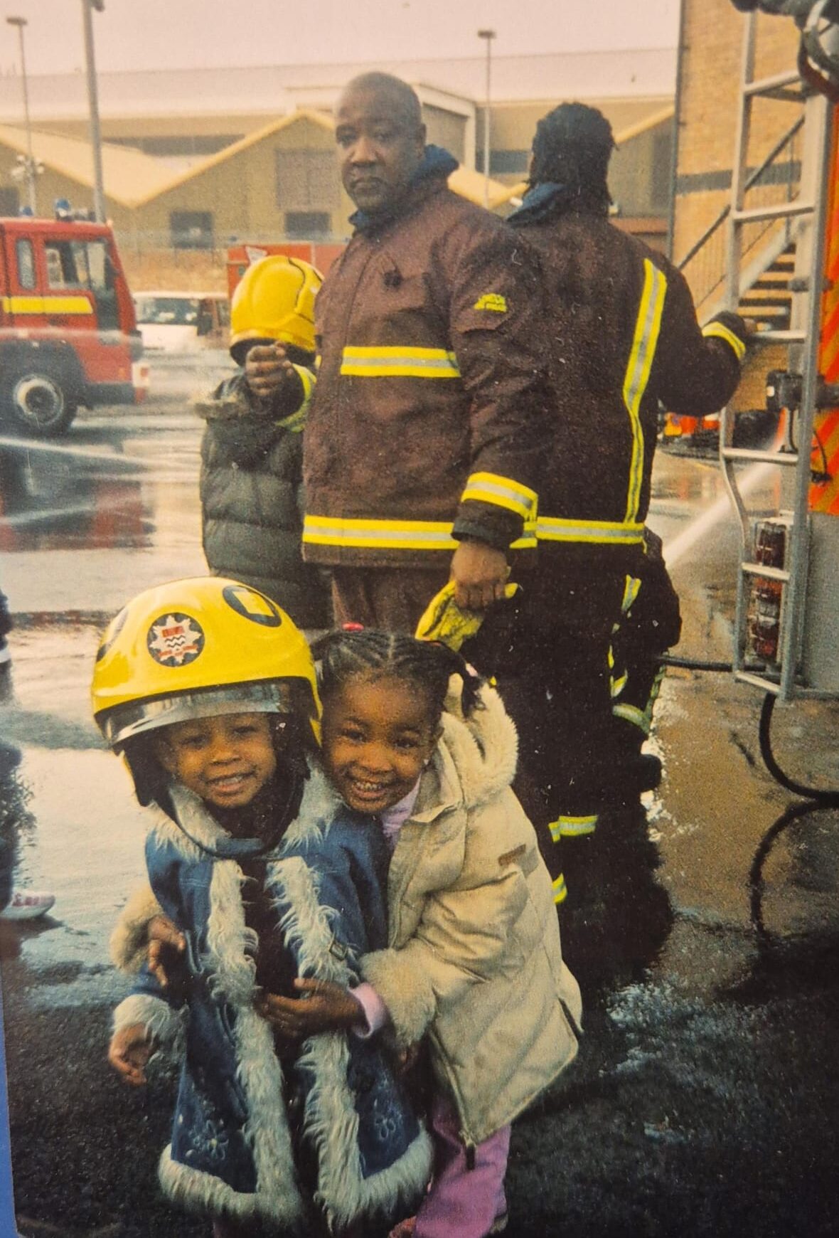 Two young girls, smiling and hugging, stand outside on a wet day. One wears a firefighter helmet. Behind them, firefighters in uniform stand near a fire truck with a ladder, and another fire truck is in the background.