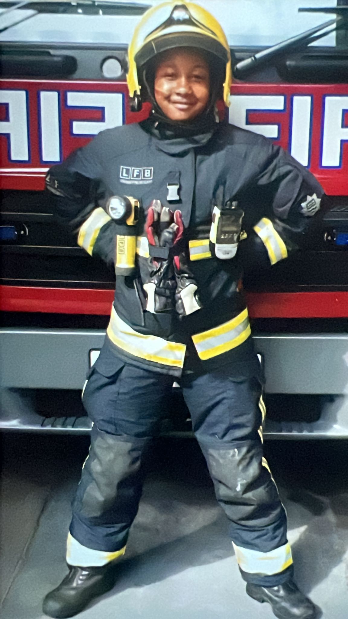 A smiling child wearing a full firefighter uniform and helmet stands in front of a fire engine, hands on hips, with firefighting tools attached to the uniform.