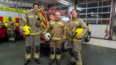 Three firefighters in full gear and holding yellow helmets stand and laugh together inside a fire station bay, with fire trucks and another firefighter visible in the background.