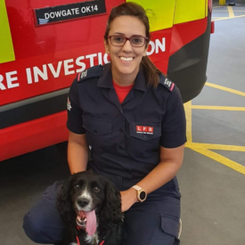 A smiling firefighter in uniform kneels beside a black and white dog in front of a red fire investigation vehicle inside a parking area.