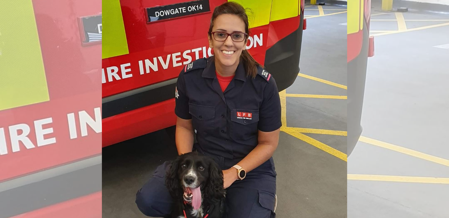 A smiling firefighter in uniform kneels beside a black and white dog in front of a red fire investigation vehicle inside a parking area.